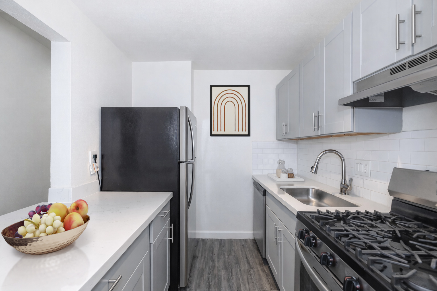 Modern kitchen featuring gray cabinets, black refrigerator, stainless steel sink, and gas stove. Countertops are white with a bowl of assorted fruits. Wall art above the counter shows a minimalist design with an arched pattern. The space is bright and neatly organized.