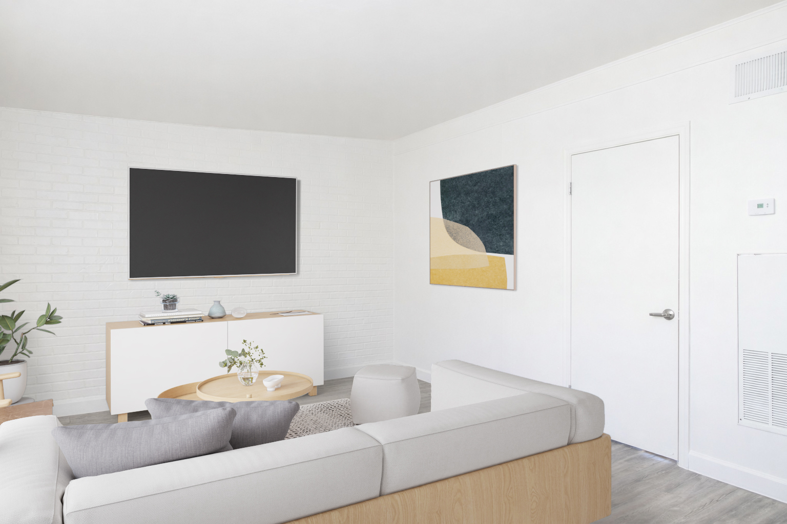Modern living room featuring a light gray sectional sofa with gray and white pillows, a round wooden coffee table, and a sleek white entertainment unit with a flat-screen TV. A minimalist wall art piece hangs above the unit, and a door is visible on the right. Natural light fills the room, creating a bright atmosphere.