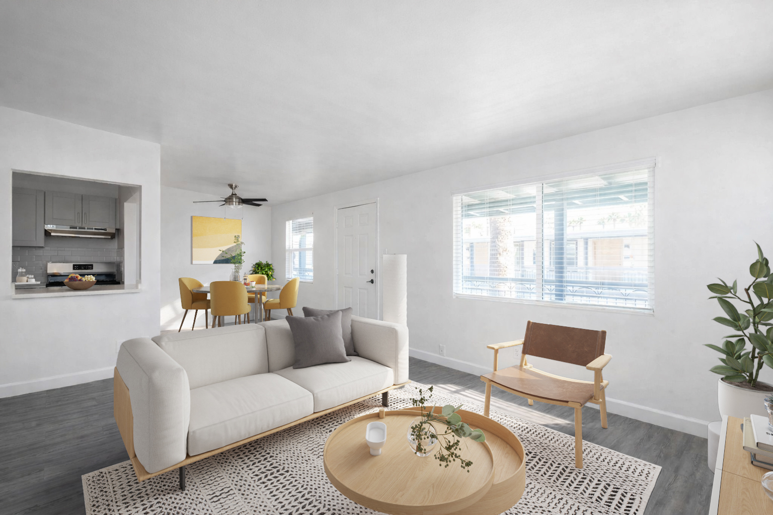 Bright and modern living room featuring a light-colored sofa with a gray cushion, a round coffee table with decorative plants, and an accent chair. A dining area with a circular table and chairs is visible in the background, along with large windows letting in natural light. The walls and flooring are neutral-toned.