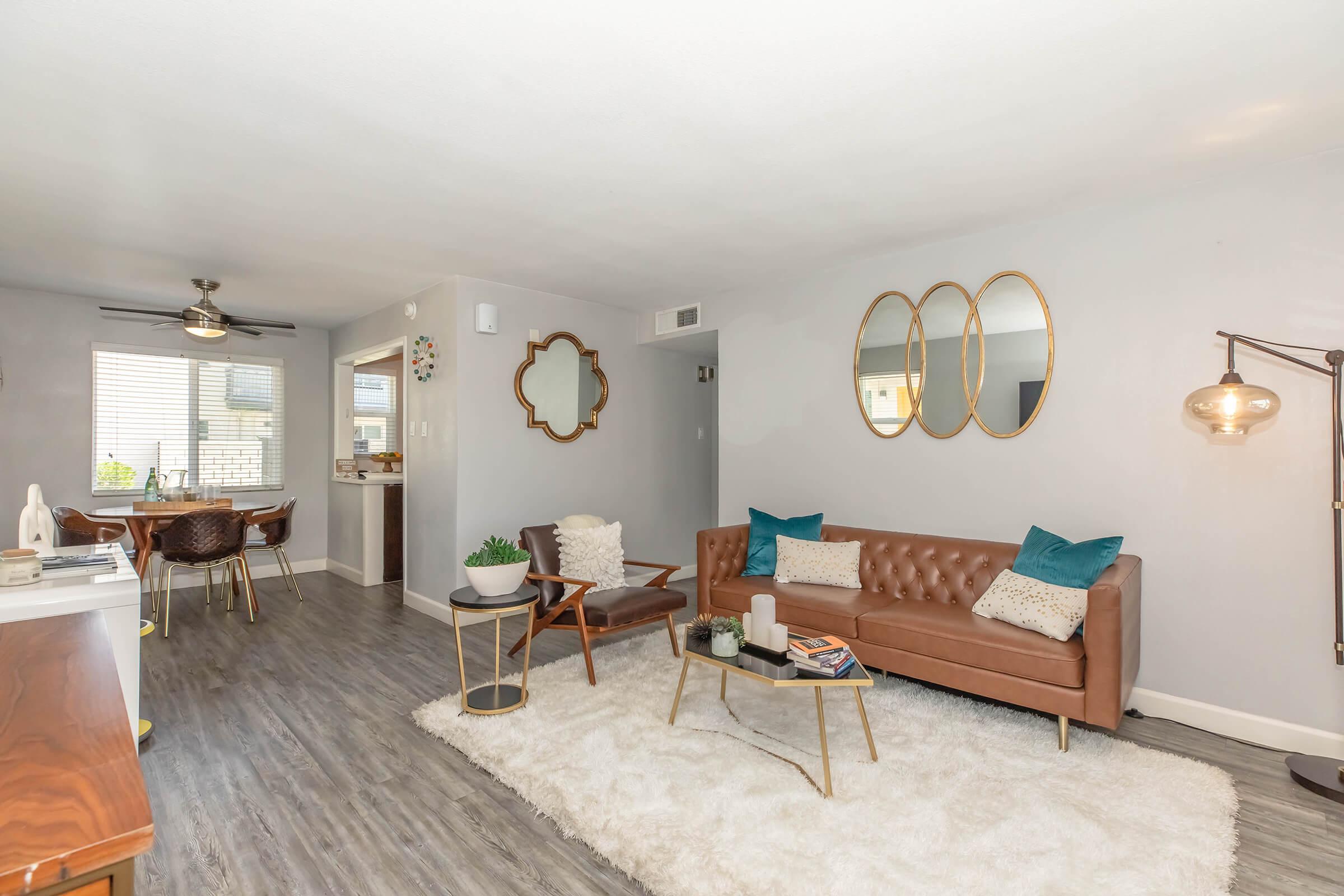 A modern living room featuring a tan leather sofa with teal and patterned cushions, a cozy white rug, a wooden coffee table, and decorative mirrors on the wall. There’s a dining area with stylish chairs in the background and a ceiling fan above, creating a warm and inviting atmosphere.