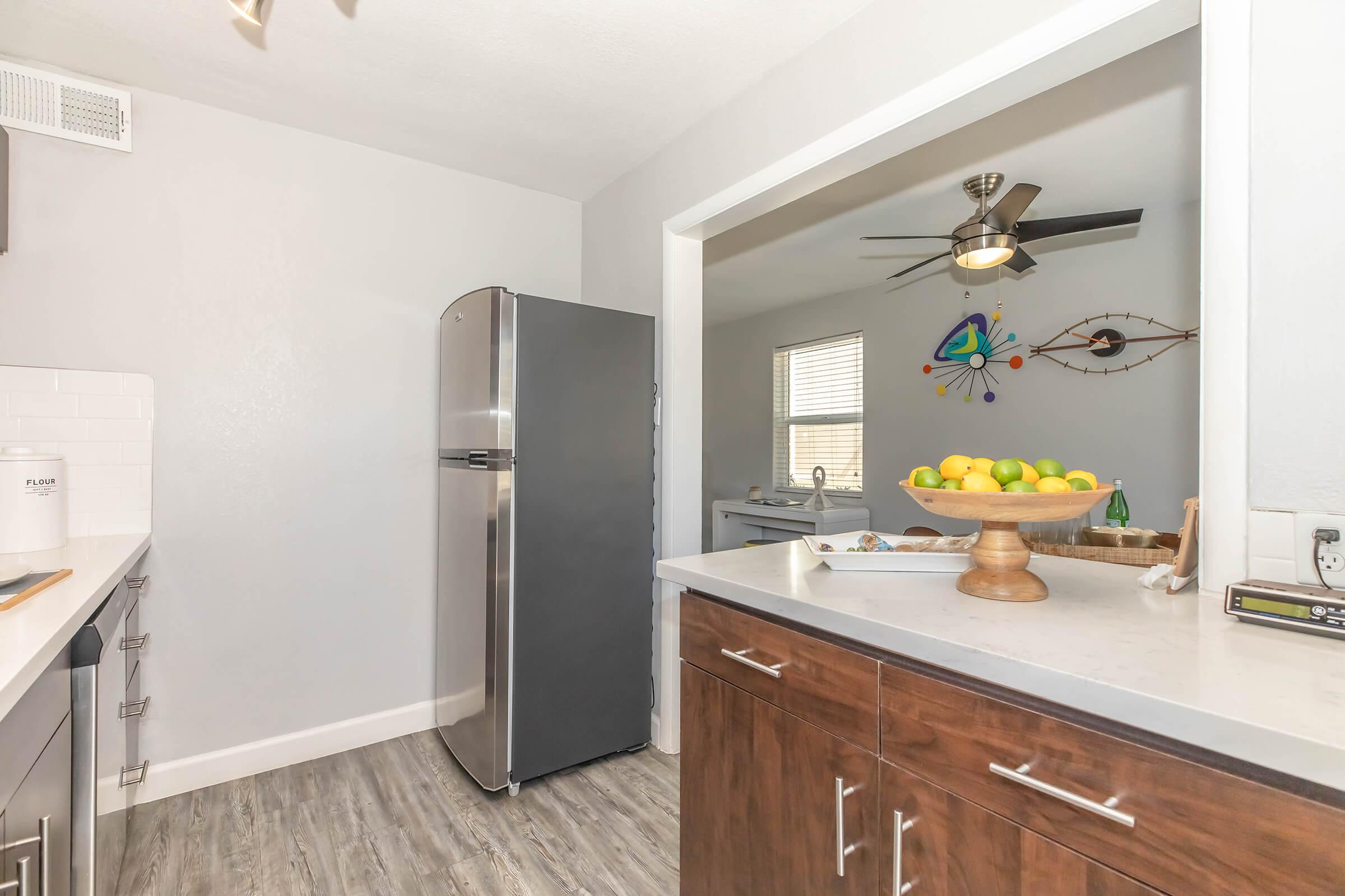Modern kitchen with stainless steel refrigerator, light gray walls, and wooden cabinetry. A bowl of colorful fruit sits on the counter, and a ceiling fan is visible. The image includes a spacious view into an adjacent room with decorative wall art and window.