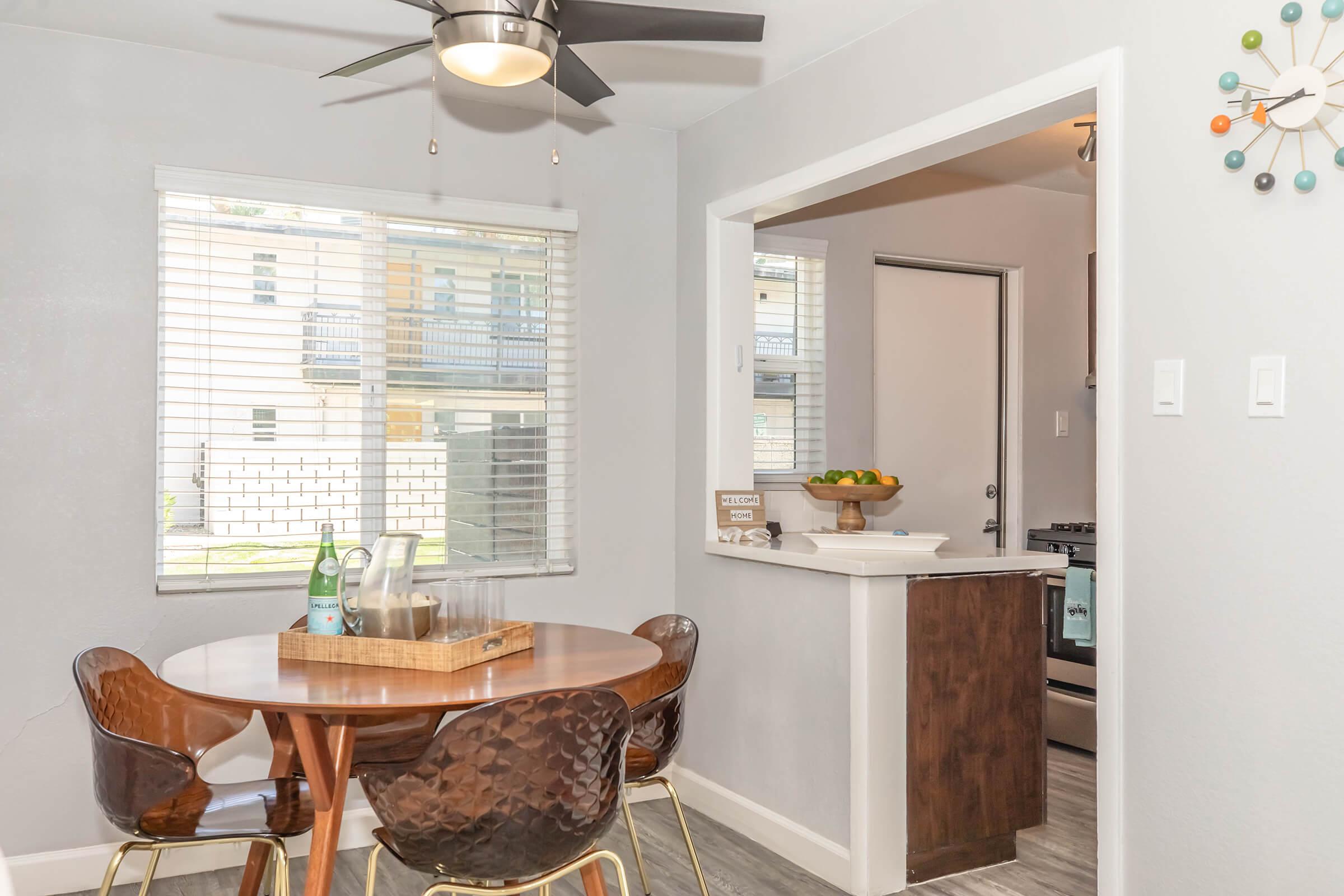 A cozy dining area featuring a round wooden table with four stylish chairs, a ceiling fan, and a bowl of fruit on the nearby counter. The walls are painted light gray, and natural light streams in through the window, illuminating the space.