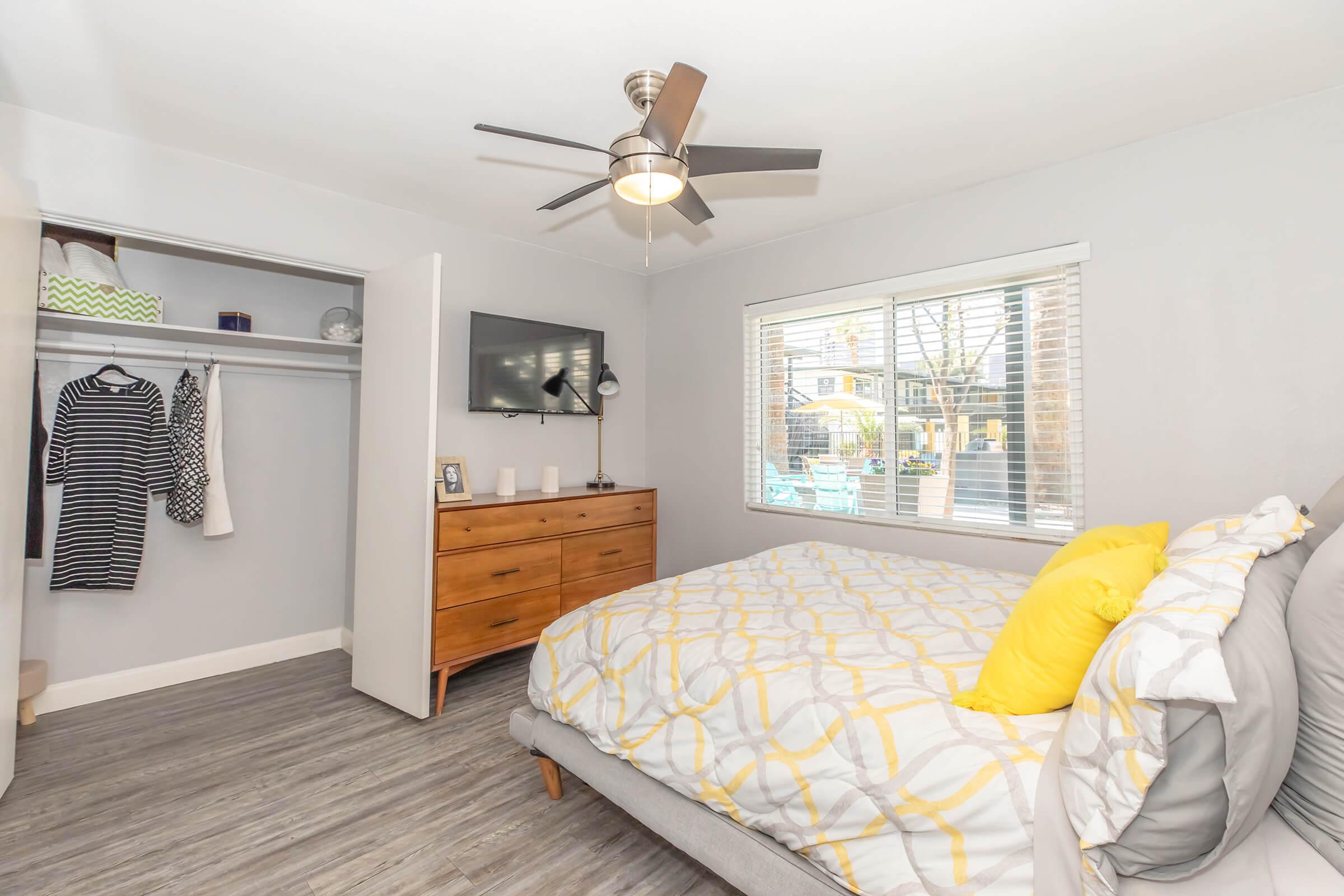 A bright, modern bedroom featuring a gray bed with a yellow accent pillow, a wooden dresser, and a flat-screen TV mounted on the wall. There’s a closet with clothes hanging inside, a large window letting in natural light, and a view of an outdoor area with patio furniture.