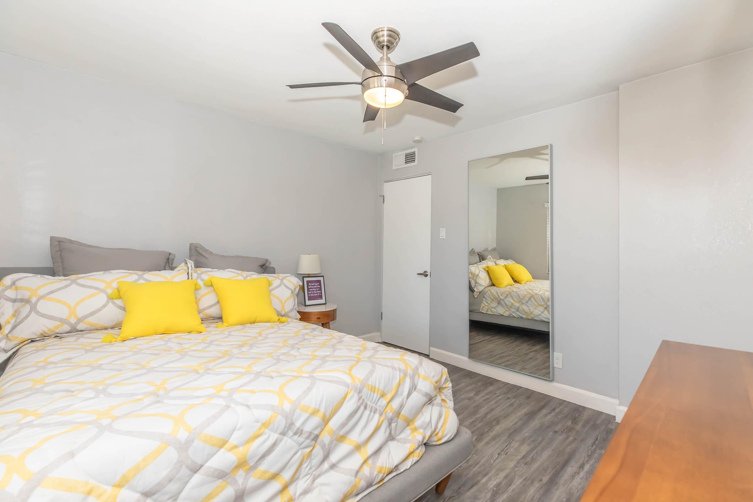 A well-lit bedroom featuring a large bed with gray and yellow patterned bedding and matching yellow pillows. There's a wooden bedside table with a lamp, a wall-mounted mirror reflecting the room, and a ceiling fan overhead. The walls are painted in a soft gray, creating a cozy atmosphere.