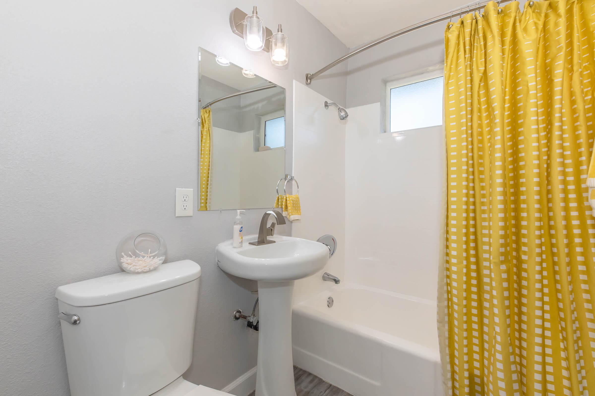 A bright and modern bathroom featuring a white toilet, a pedestal sink, and a bathtub with a shower. The space is adorned with a cheerful yellow shower curtain with white polka dots, and there is a round mirror above the sink. Soft natural light streams in through a small window.