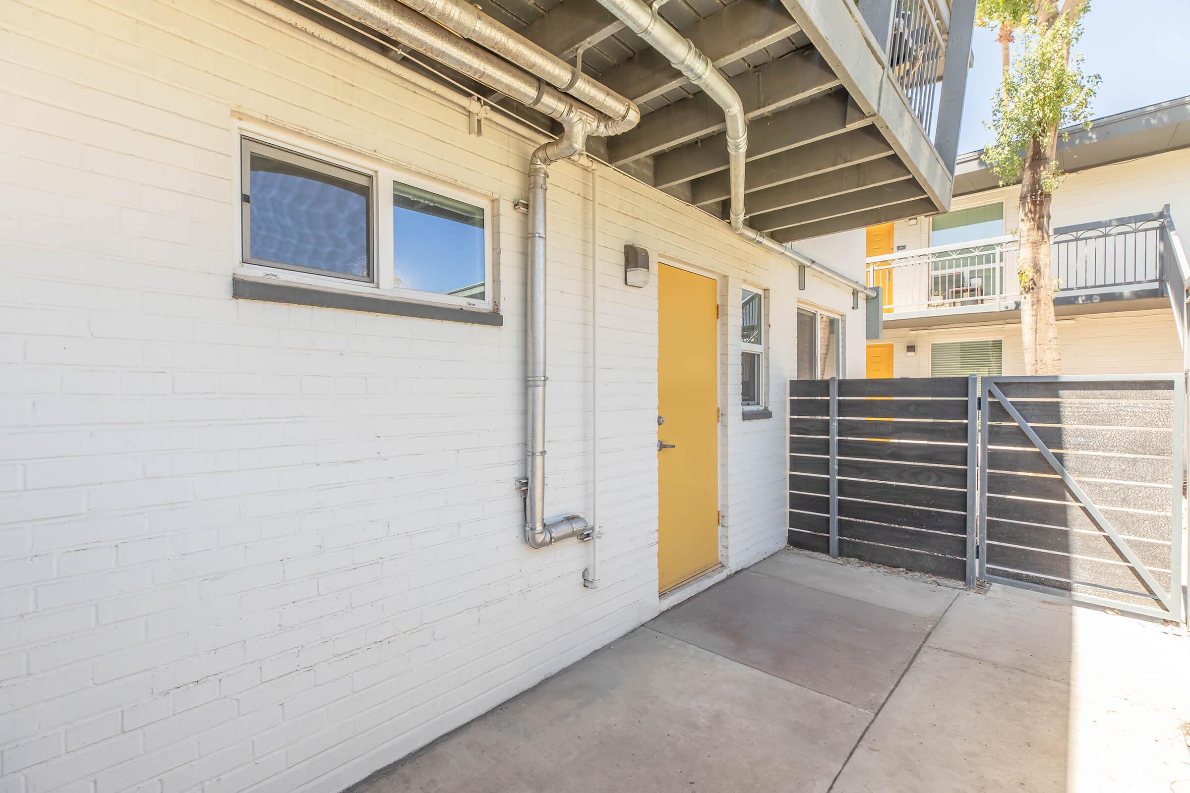 A modern entrance featuring a yellow door set against a white brick wall. There is a metal railing overhead and a gray fence to the right, partially enclosing a small pathway that leads to the door. Sunlight casts shadows on the concrete floor, enhancing the contemporary design of the space.
