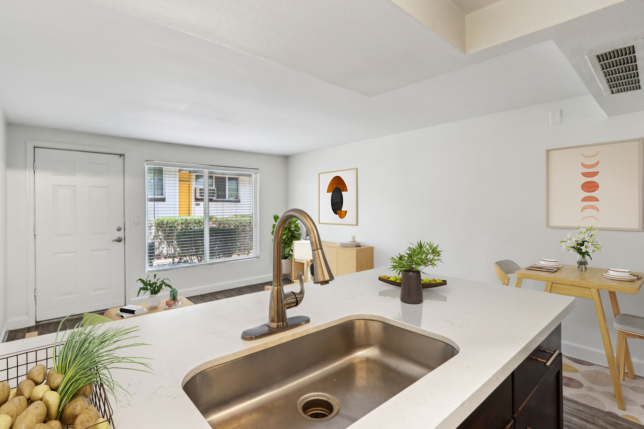 A modern kitchen with a view of a living area. The kitchen features a stainless steel sink and a countertop with decorative plants. In the background, there's a dining table set with dishes and a stylish wall art. Natural light streams through a window, creating a bright, inviting atmosphere.