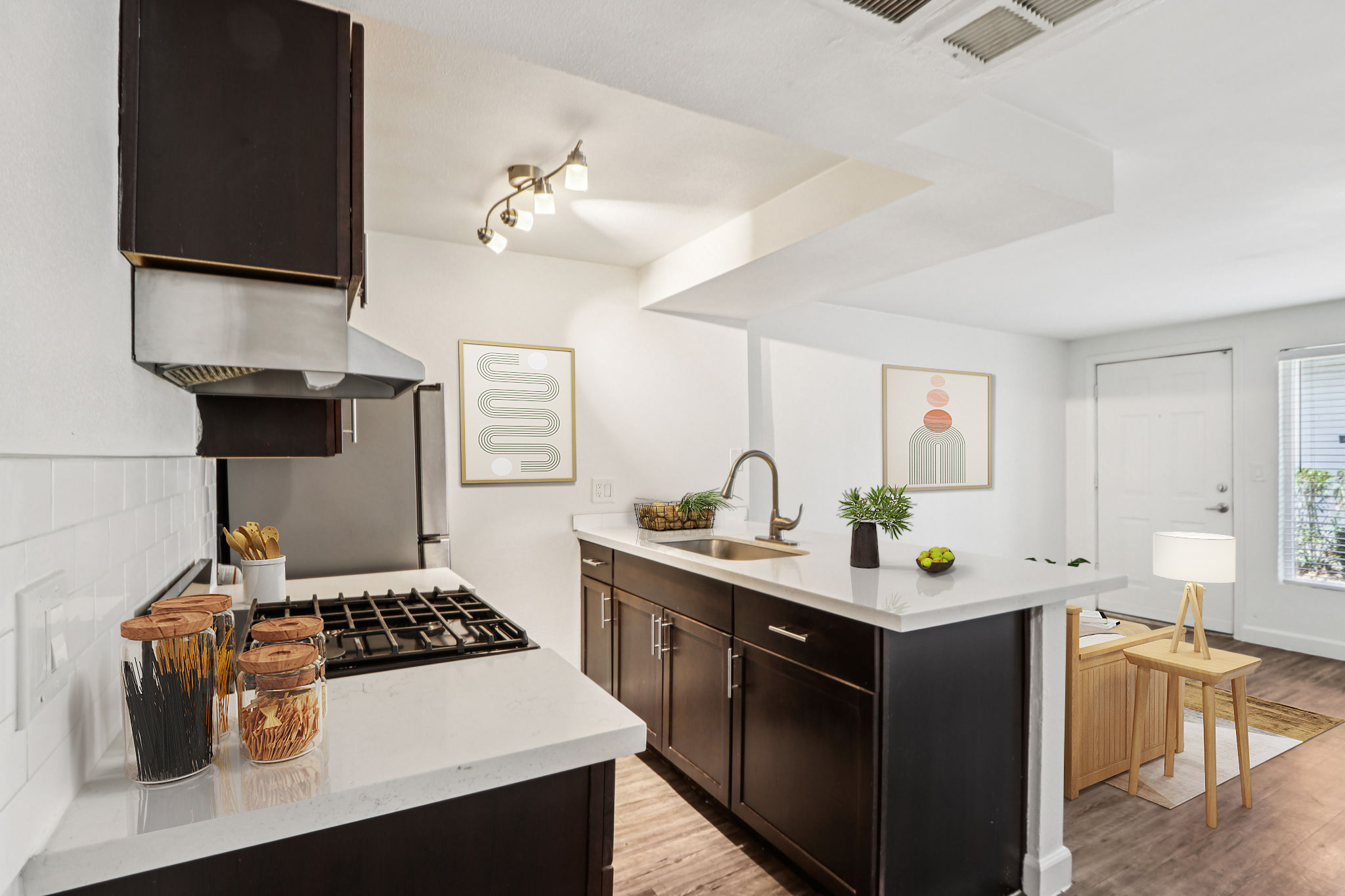 Modern kitchen featuring dark cabinetry, a stainless steel stove and hood, and a white countertop. The space includes decorative elements like plants and framed artwork on the walls, with natural light illuminating the room from a doorway.