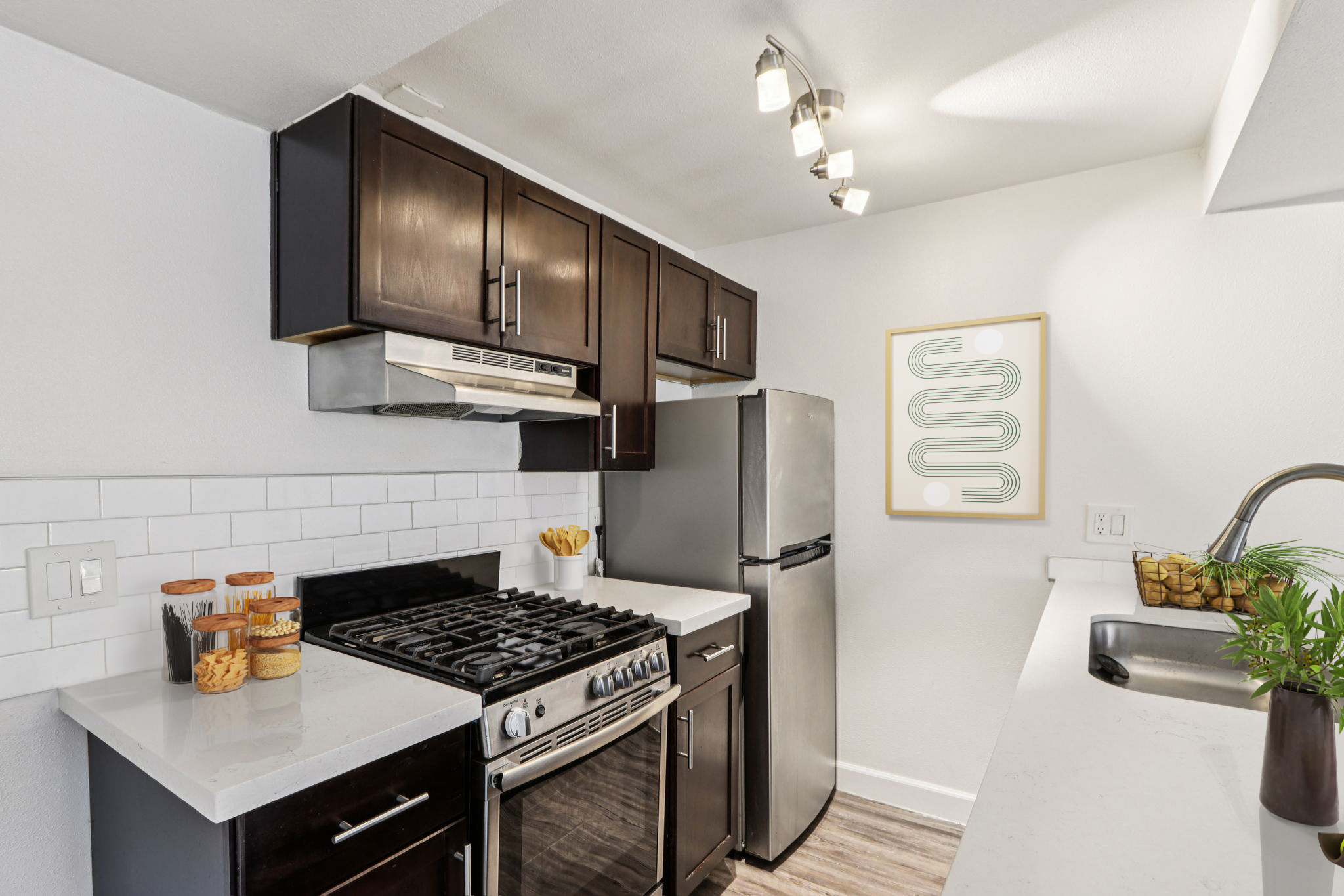 Modern kitchen featuring dark wood cabinets, stainless steel appliances, and a white countertop. A gas stove is in the foreground, with a refrigerator nearby. Decorative jars are placed on the counter, and a simple framed wall art piece adds a touch of style. Well-lit with track lighting.