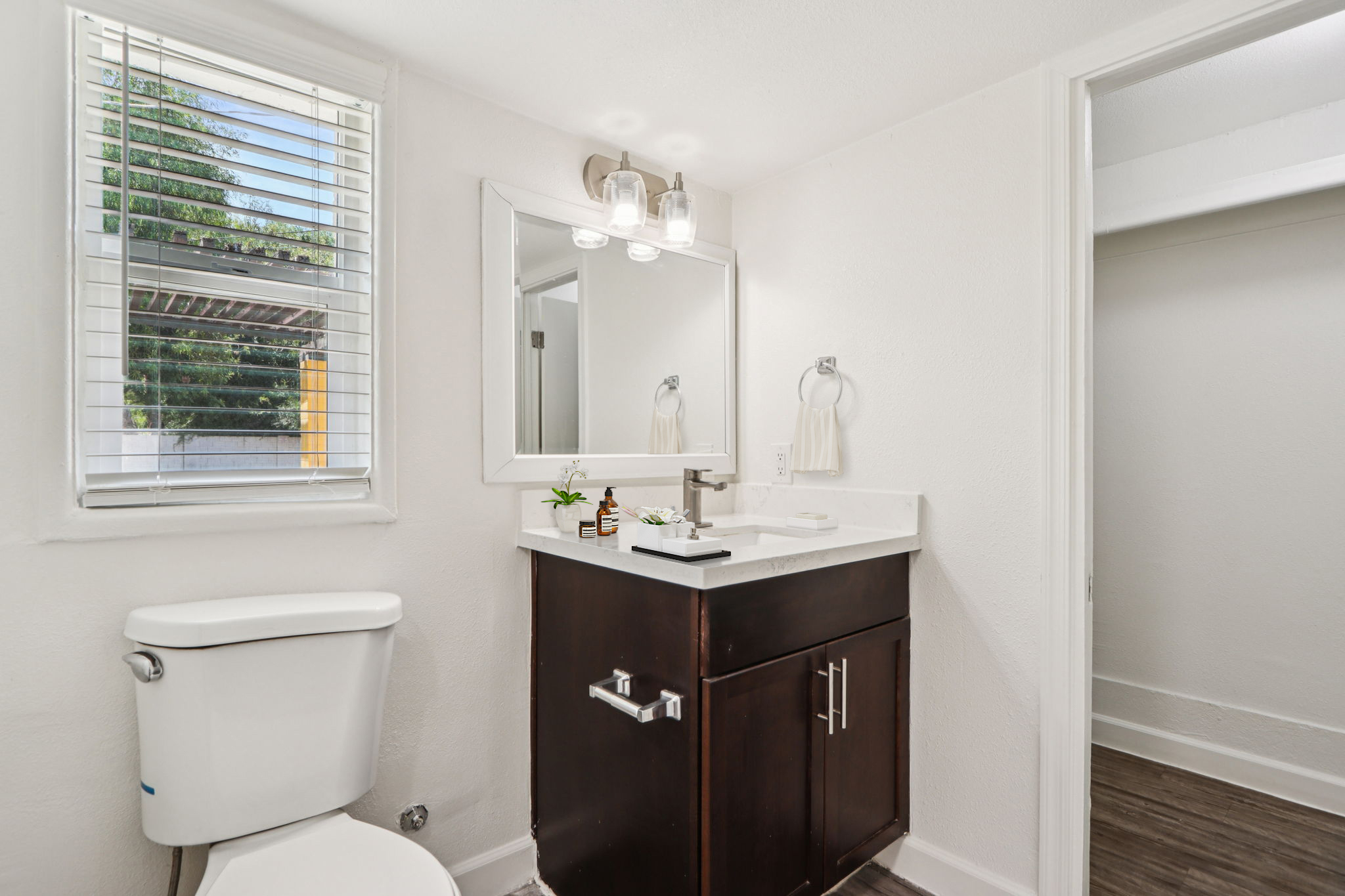 A modern bathroom featuring a white toilet, a dark wood vanity with a sink, a wall-mounted mirror, and stylish light fixtures. Natural light filters in through a window with blinds, illuminating the clean and minimalist design of the space. The floor is finished with wood-like flooring.