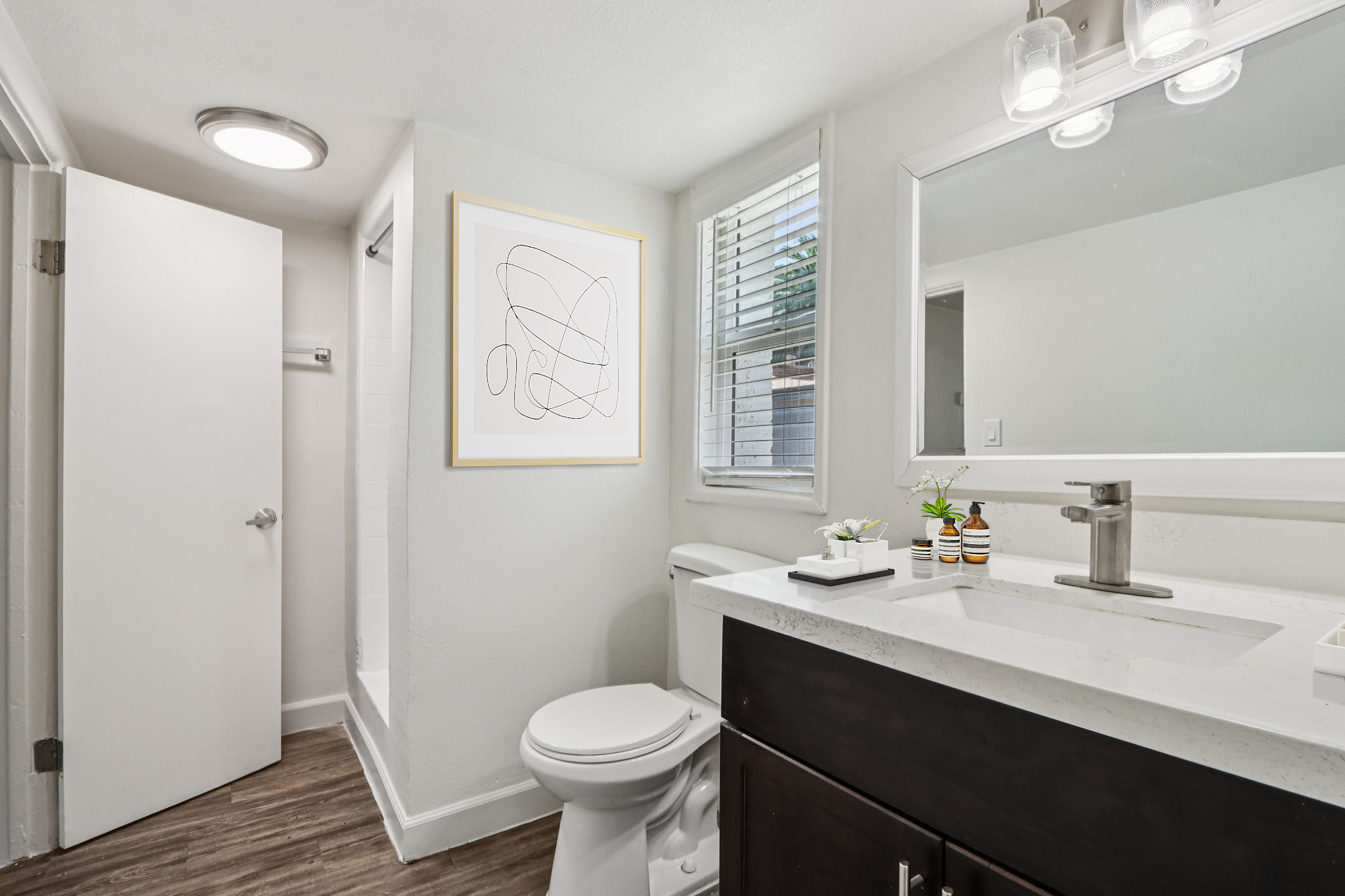 A modern bathroom featuring a white toilet, a sleek vanity with a sink, and a large mirror. There is a framed abstract art piece on the wall, stylish plants on the countertop, and a shower area visible in the background. The color scheme is light and airy, with natural light coming through a window.