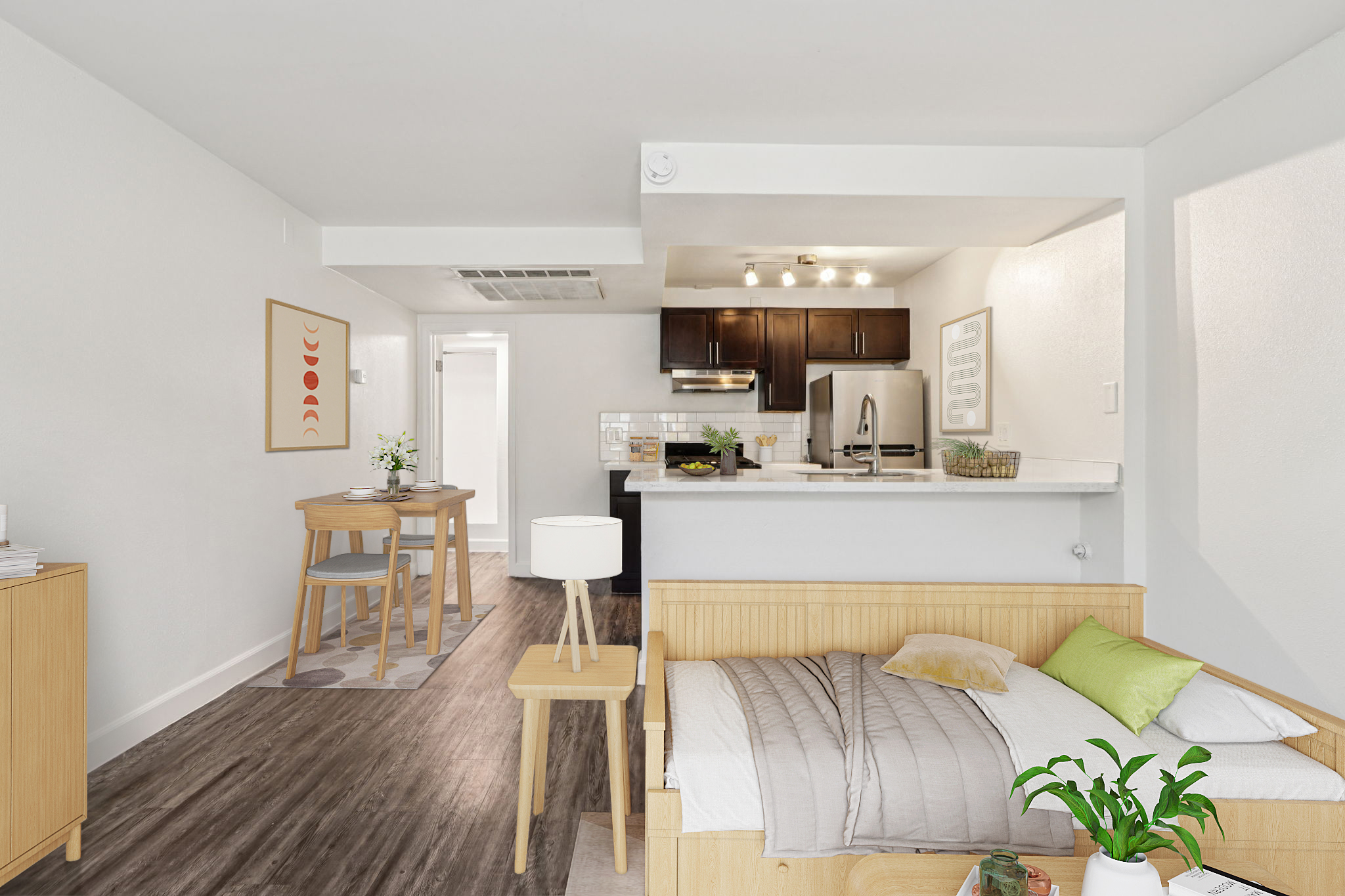 A modern, open-concept living space featuring a wooden bed with white bedding and a decorative pillow, a light wood dining table with two chairs, and a styled kitchen area with dark cabinetry and stainless steel appliances. The room is bright with neutral walls and a plant adding a touch of greenery.