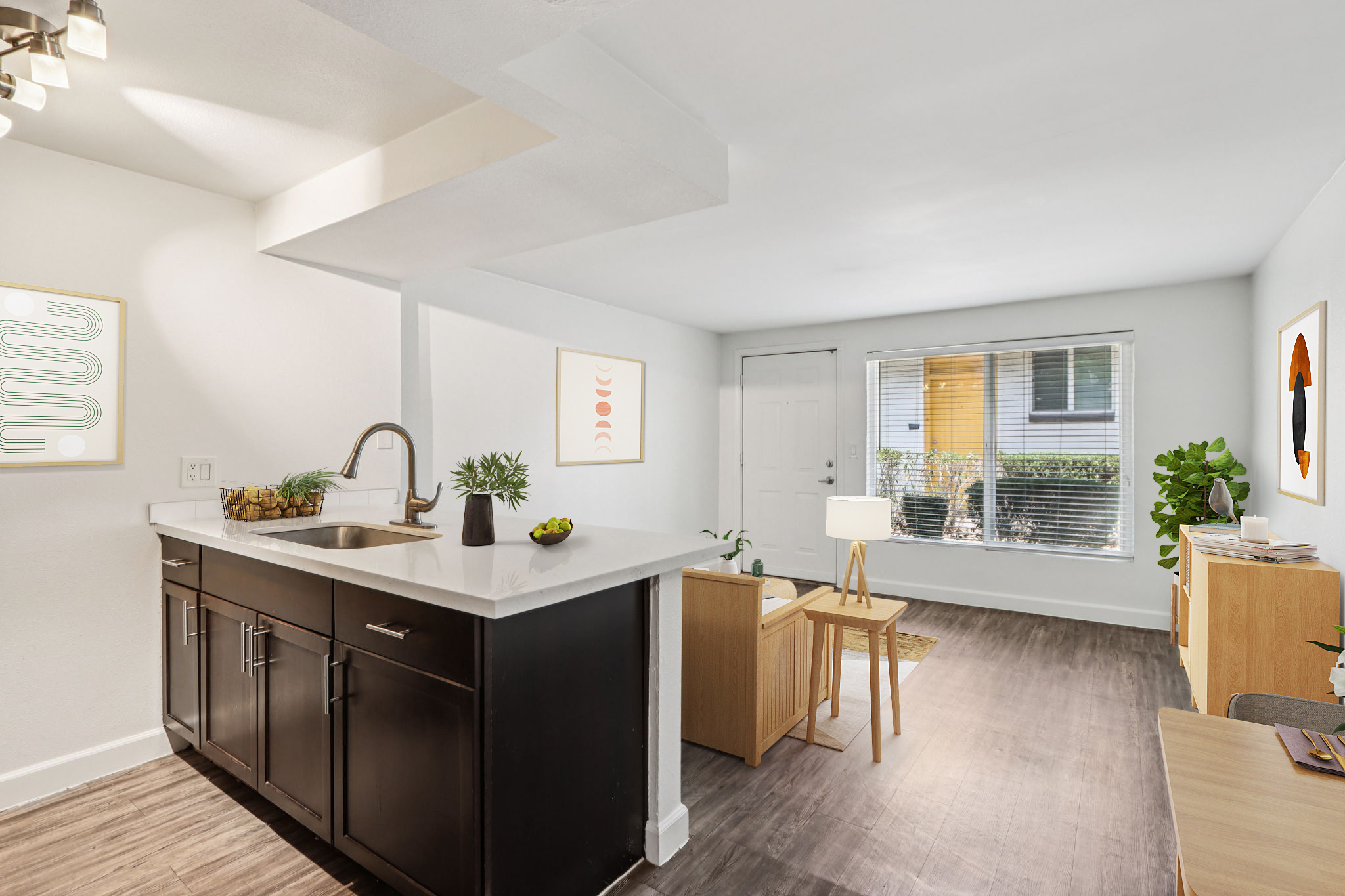 A modern living space featuring a kitchenette with a dark cabinet and white countertop, a small dining area with a lamp, plants, and artwork on the walls. Natural light floods the room through a window, showcasing a cozy atmosphere with neutral decor and minimalist furnishings.