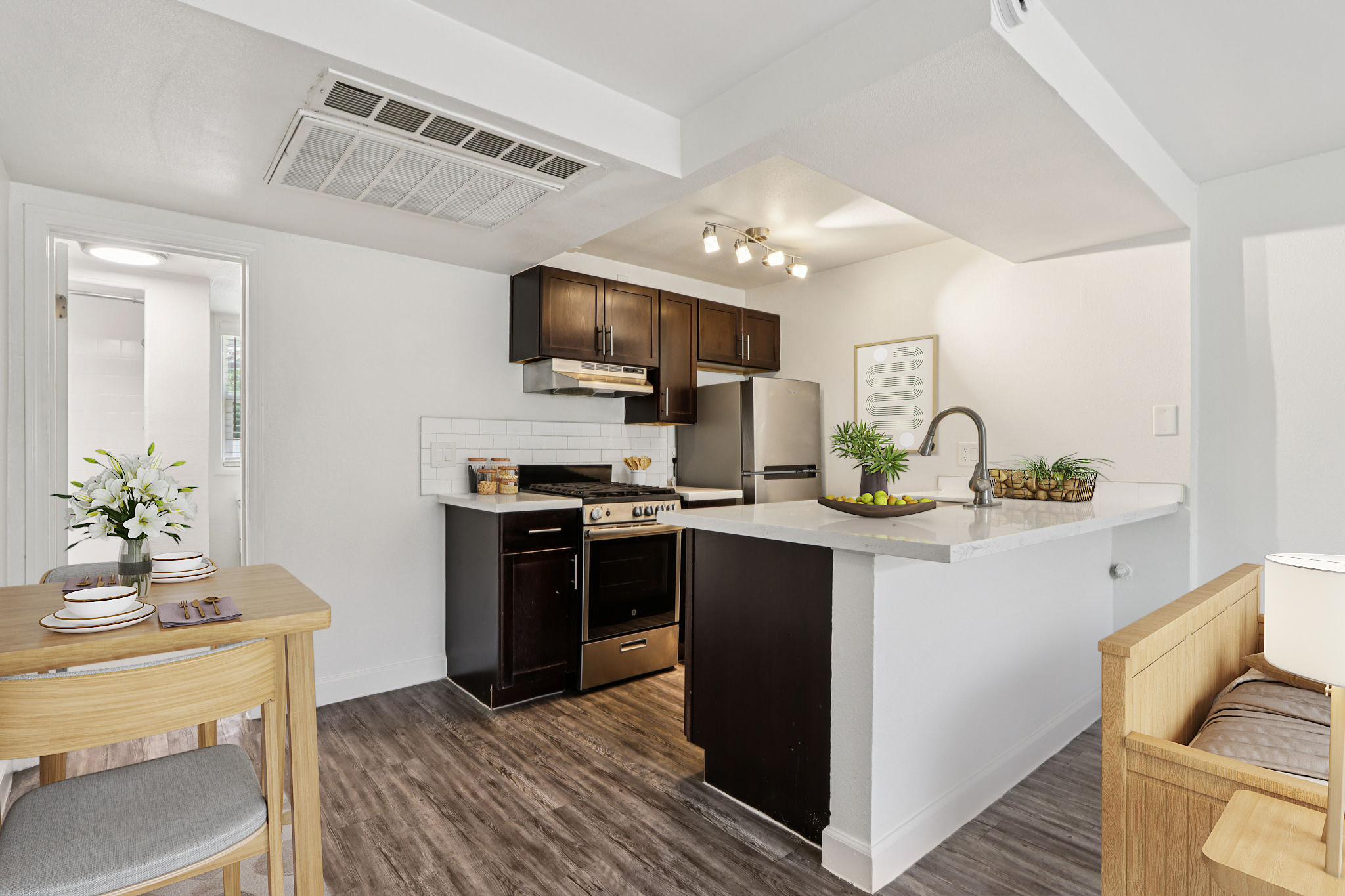 A modern kitchen featuring dark wood cabinets, stainless steel appliances, and a white countertop. A dining table with place settings is in the foreground, and potted plants add a touch of greenery. The space is bright and well-lit, creating an inviting atmosphere.