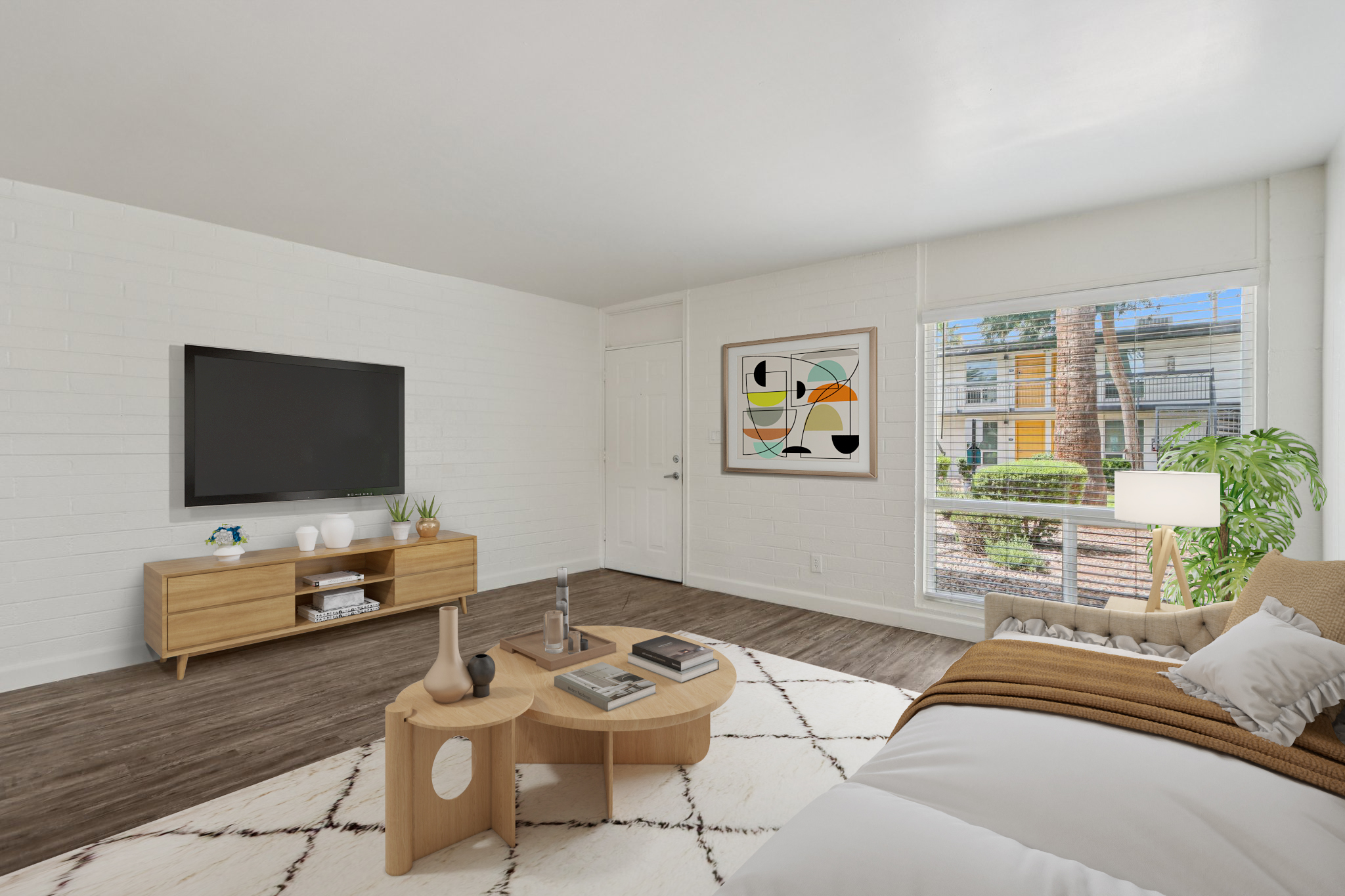 A modern living room featuring a beige sofa with a white throw blanket, a round coffee table with a decorative vase, and a television mounted on a white brick wall. There’s a large window providing natural light, a potted plant, and an abstract artwork on the wall, set in a bright, open space.