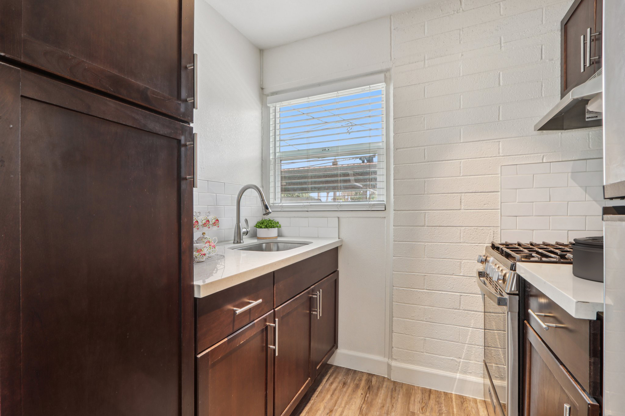 A modern kitchen featuring dark wooden cabinets, a stainless steel gas stove, and a countertop with a sink. The backsplash is white tile, and there's a window allowing natural light and a small potted plant on the counter. The floor has a wood-like finish, creating a warm ambiance.