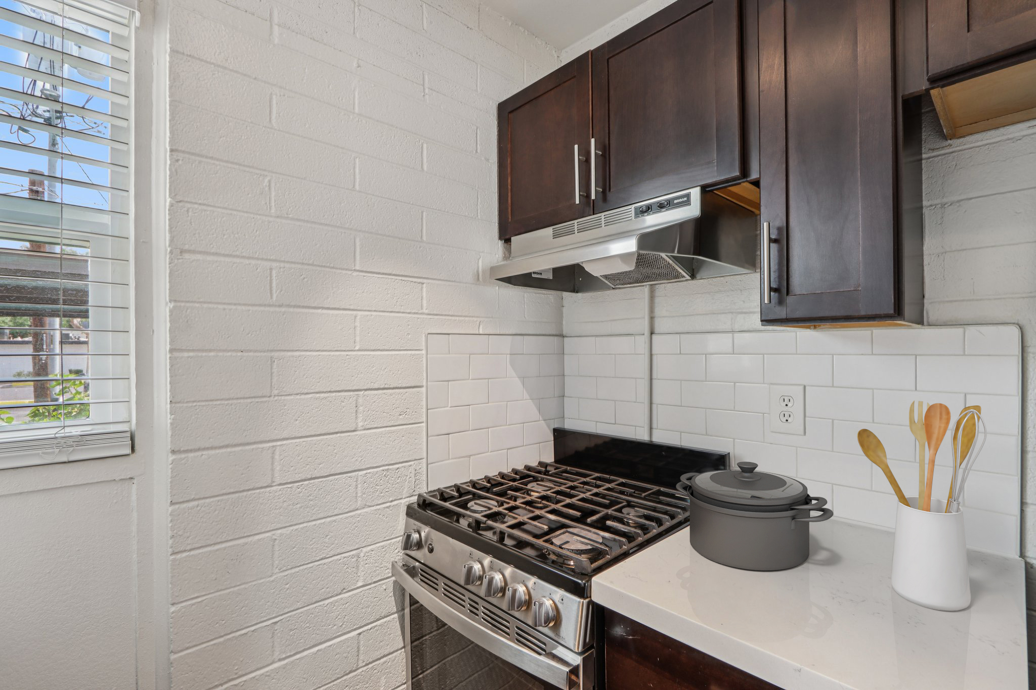A modern kitchen featuring dark wooden cabinets, white brick walls, and a gas stove with a stainless steel range hood. On the countertop, there is a black cooking pot and wooden utensils in a container. A window allows natural light to enter, enhancing the bright and functional atmosphere.