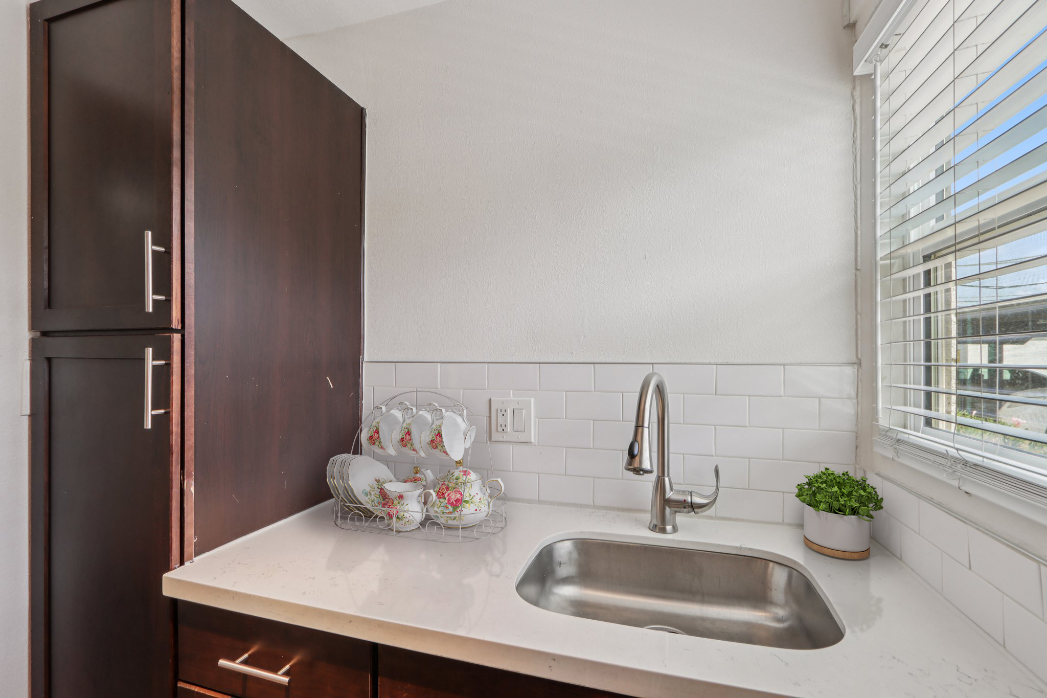 A small kitchen area featuring a stainless steel sink, white countertop, and dark wood cabinets. A wire rack holds decorative teacups beside a small potted plant, with natural light streaming in through a window with white blinds.