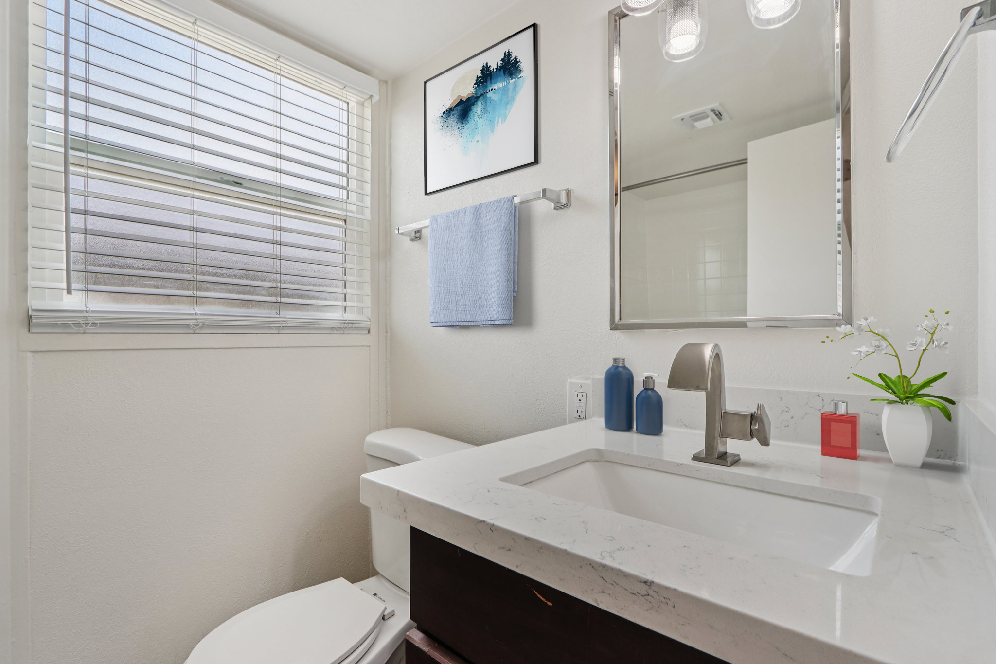 A modern bathroom featuring a bright atmosphere, with a white sink and dark wood cabinets. There is a mirror above the sink, a blue towel hanging on a rack, and a piece of art on the wall. Natural light enters through a window with blinds, enhancing the minimalist decor.