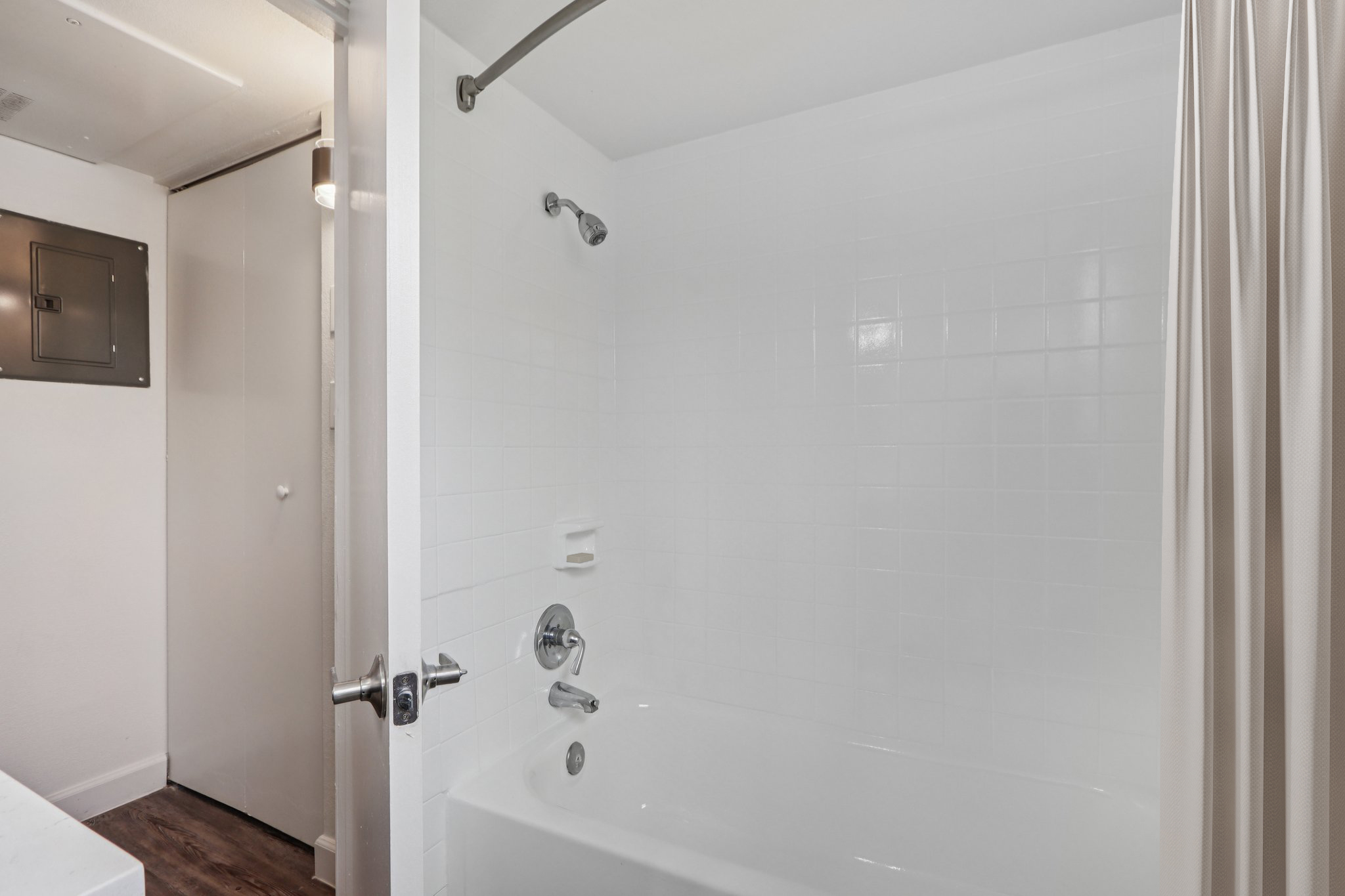 A clean, modern bathroom featuring a white bathtub and shower with a chrome faucet. The walls are tiled in white, and there's a shower curtain on one side. A door leads to another room, and the space is well-lit, creating a bright and inviting atmosphere.