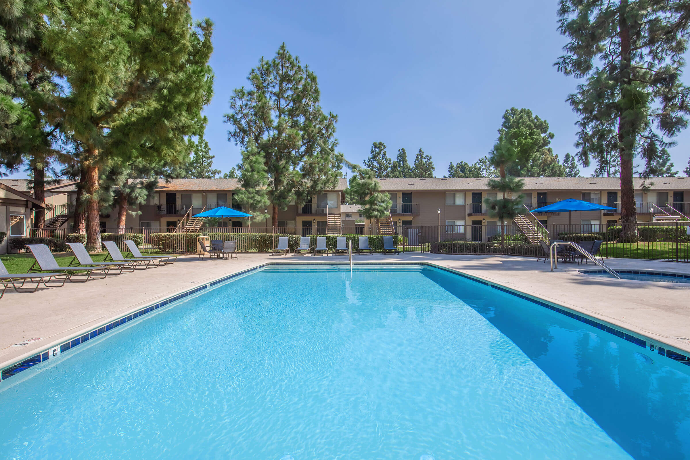 A clear swimming pool surrounded by lounge chairs, with blue umbrellas providing shade. In the background, there are two-story apartment buildings framed by tall trees, under a bright blue sky.