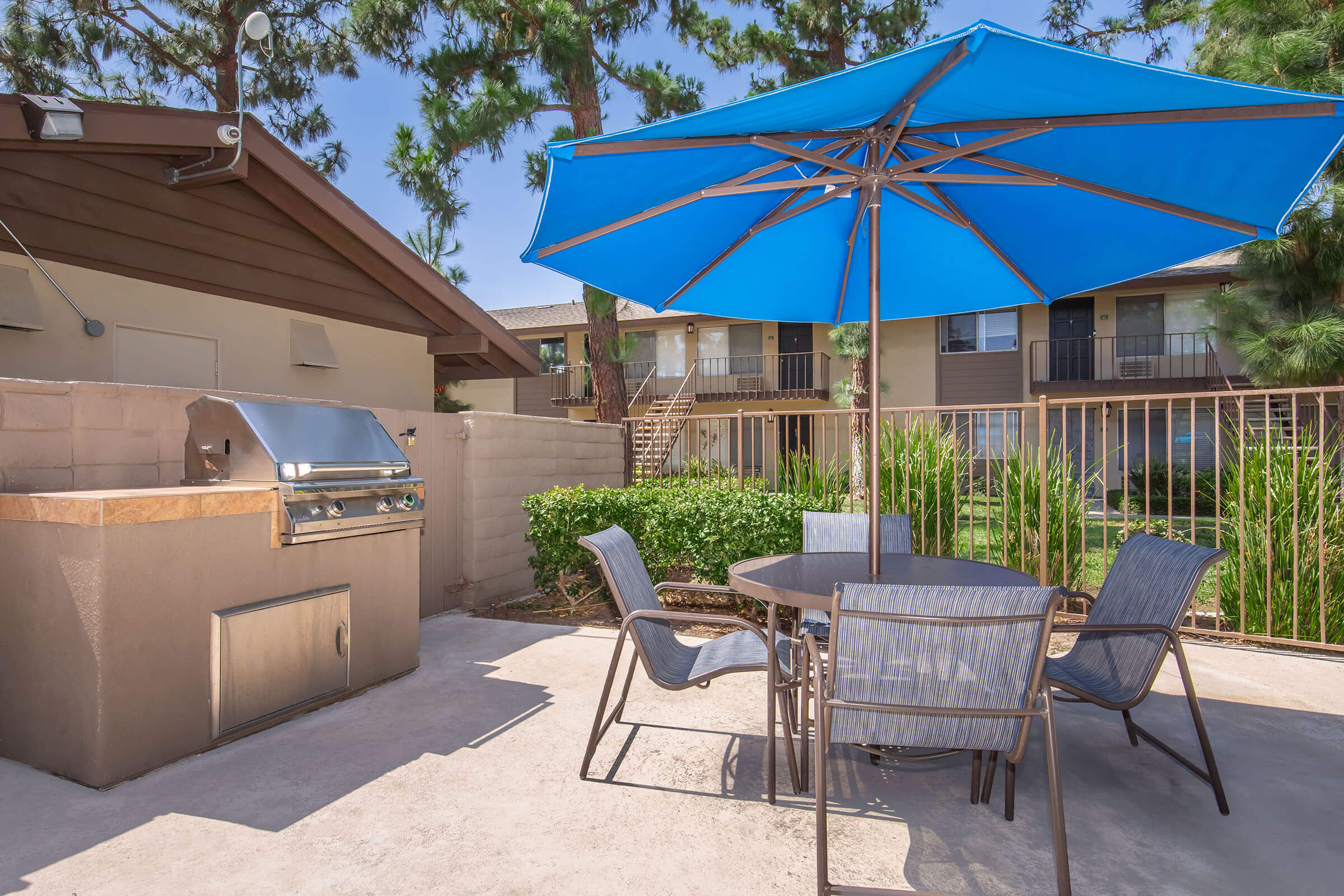 A patio area with a blue umbrella over a round table and chairs, adjacent to a barbecue grill. The background features a low wall, lush greenery, and multi-unit buildings with balconies, all under a clear blue sky.