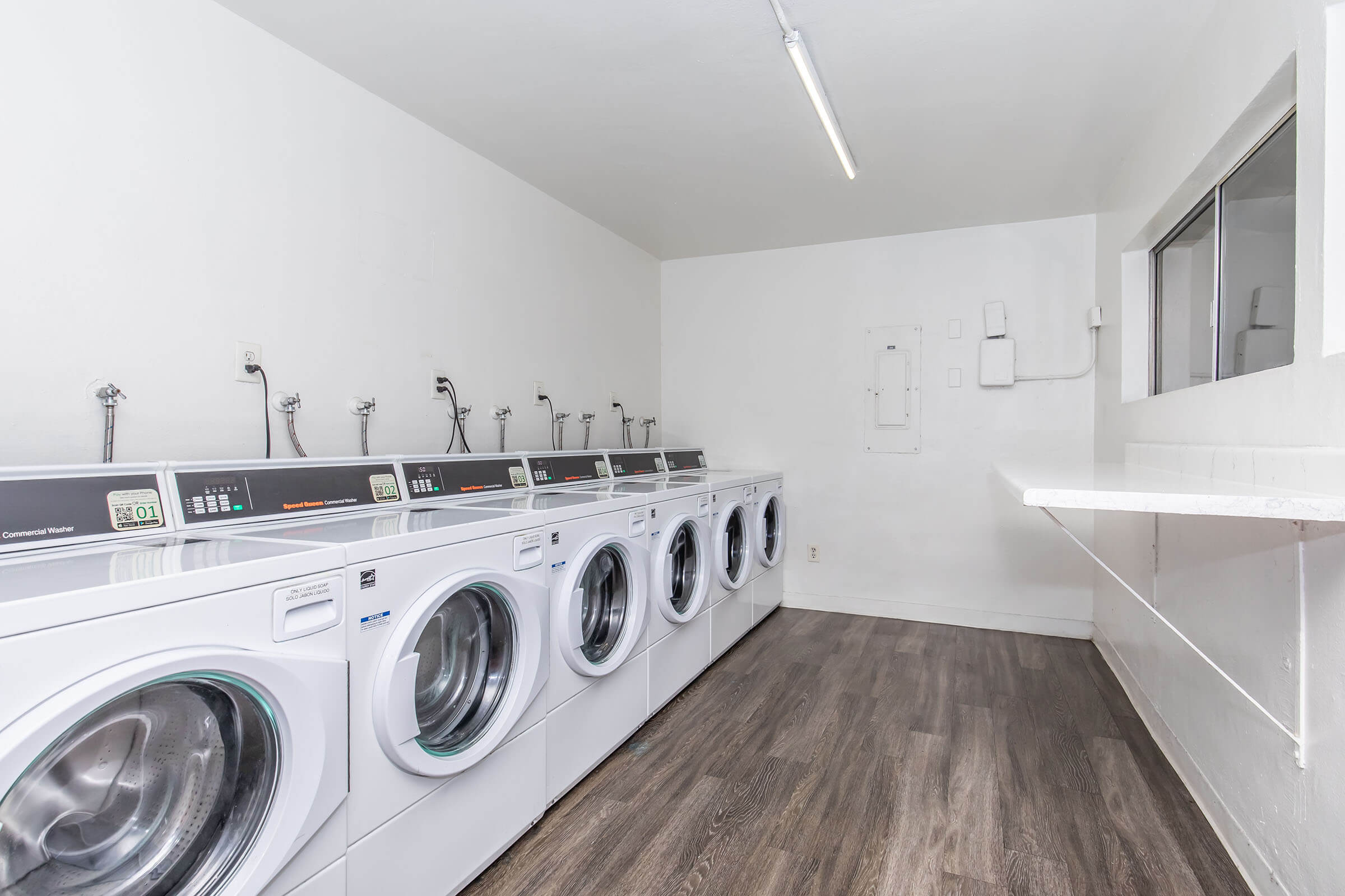 A modern laundromat with several washing machines lined up against a white wall. The machines feature digital displays and stainless steel doors. There is a counter along one side and a window that allows natural light into the space. The floor is a wooden laminate, creating a clean and bright environment.
