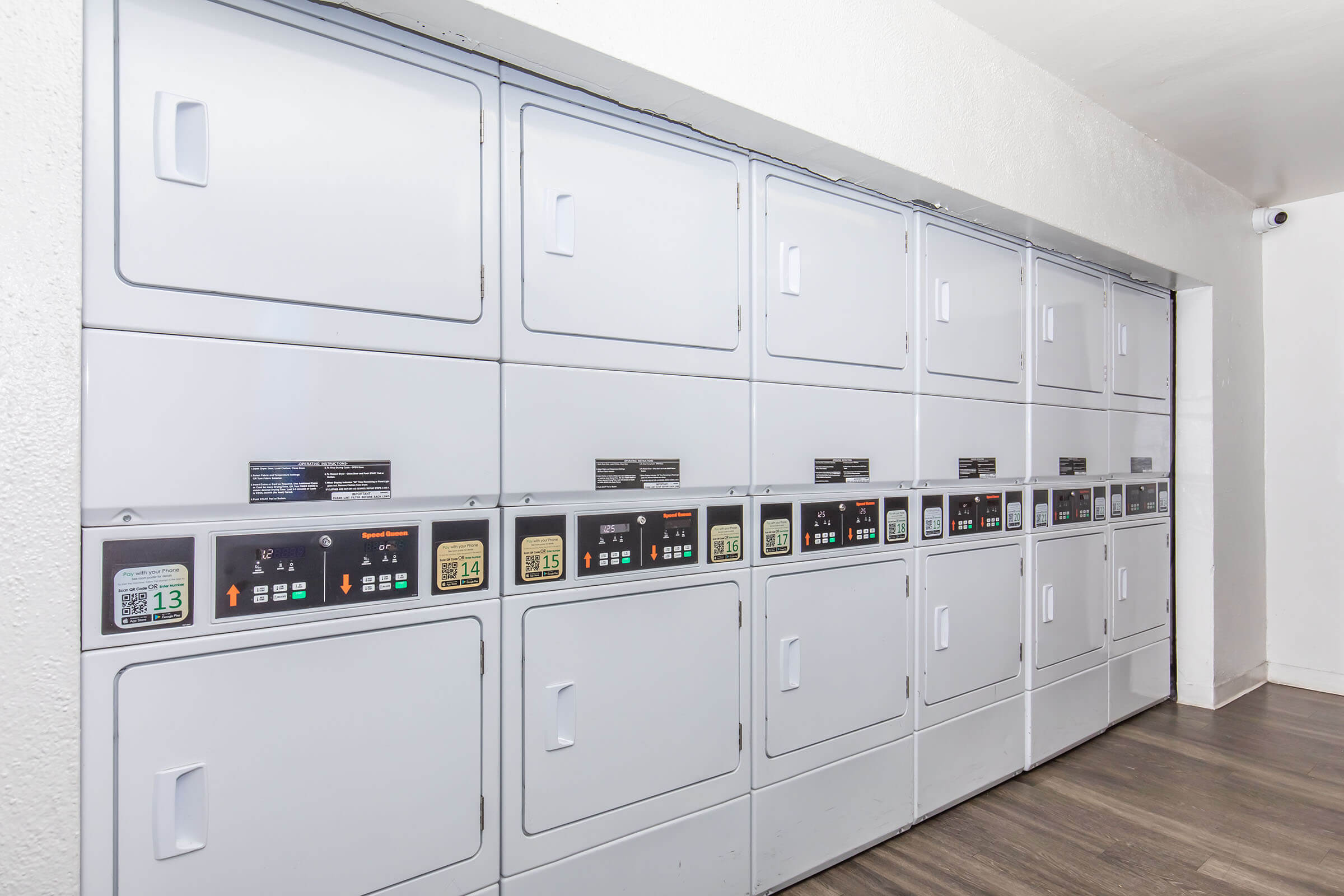 A row of modern self-service laundry machines in a clean, well-lit room. The machines feature digital displays with various buttons and indicators for selecting wash cycles. The interior is simple and minimalistic, with white walls and a wooden floor, creating a tidy and organized environment.
