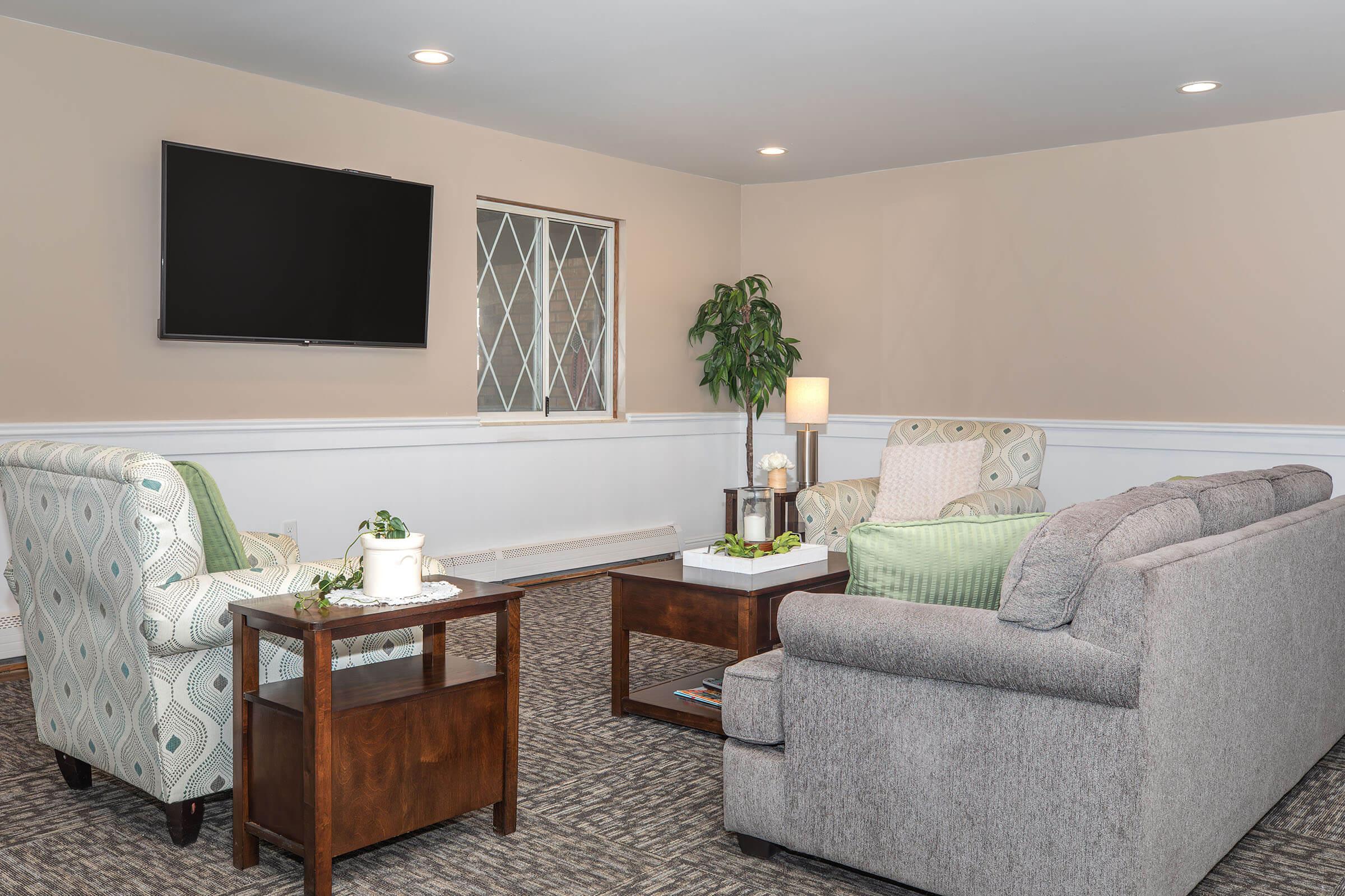 A cozy living area featuring two comfortable sofas, an armchair, and a coffee table. A flat-screen TV is mounted on the wall, and a potted plant adds a touch of greenery. Soft lighting creates a warm atmosphere, making it an inviting space for relaxation and conversation.