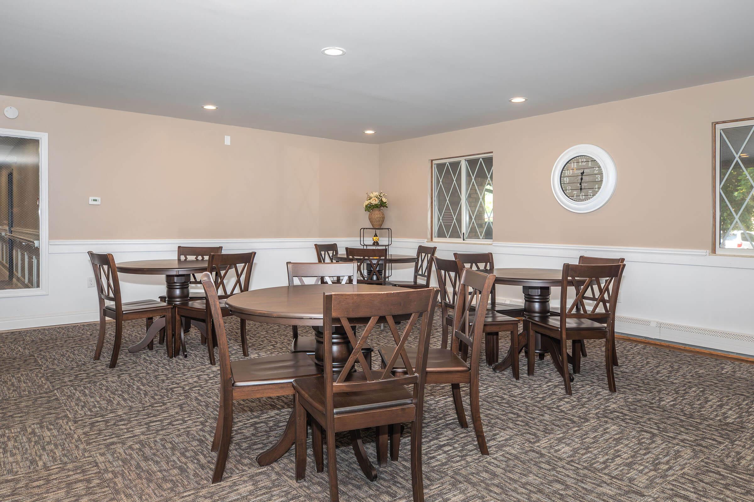 A well-lit dining area featuring several wooden tables with matching chairs, arranged neatly on a textured carpet. The walls are painted a soft beige, with windows allowing natural light, and a decorative round mirror is visible on one wall. A small vase with flowers adds a touch of decor.
