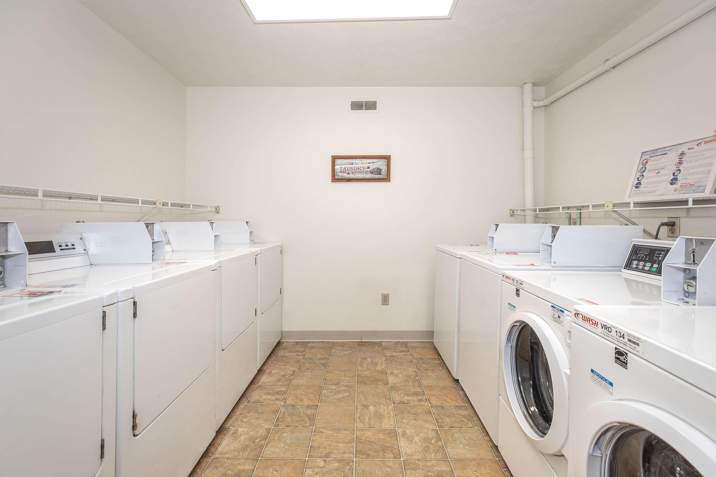 A clean and bright laundry room with several white washing machines and dryers arranged along the walls. The floor features tiled flooring, and there is a small framed picture on the wall. Soft lighting comes from a ceiling fixture, creating an inviting atmosphere.