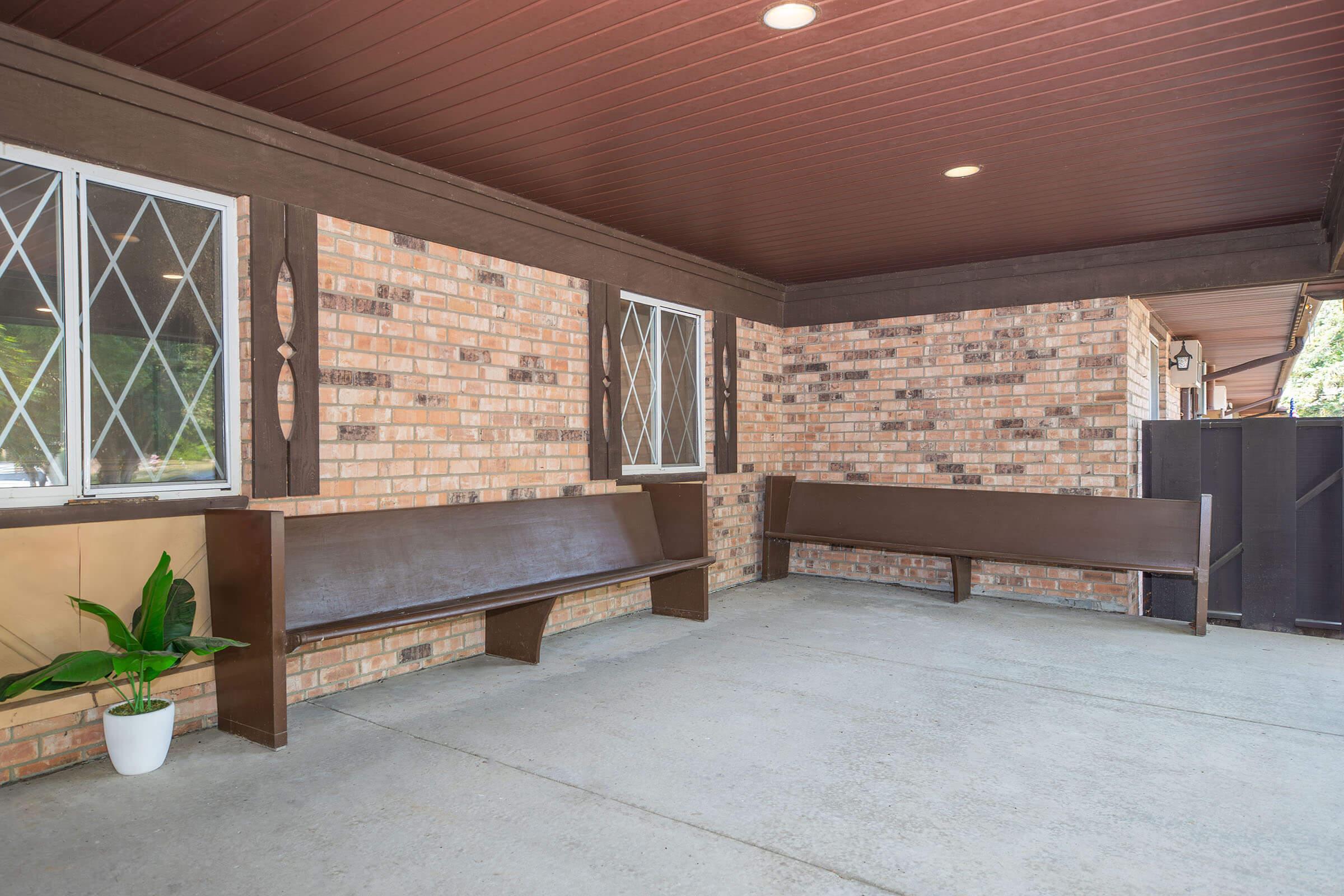 An outdoor seating area featuring two wooden benches against a brick wall, with a potted green plant on the left. The ceiling is finished in dark wood, and there are two windows with diamond-patterned panes allowing natural light into the space. The floor is a plain concrete surface.