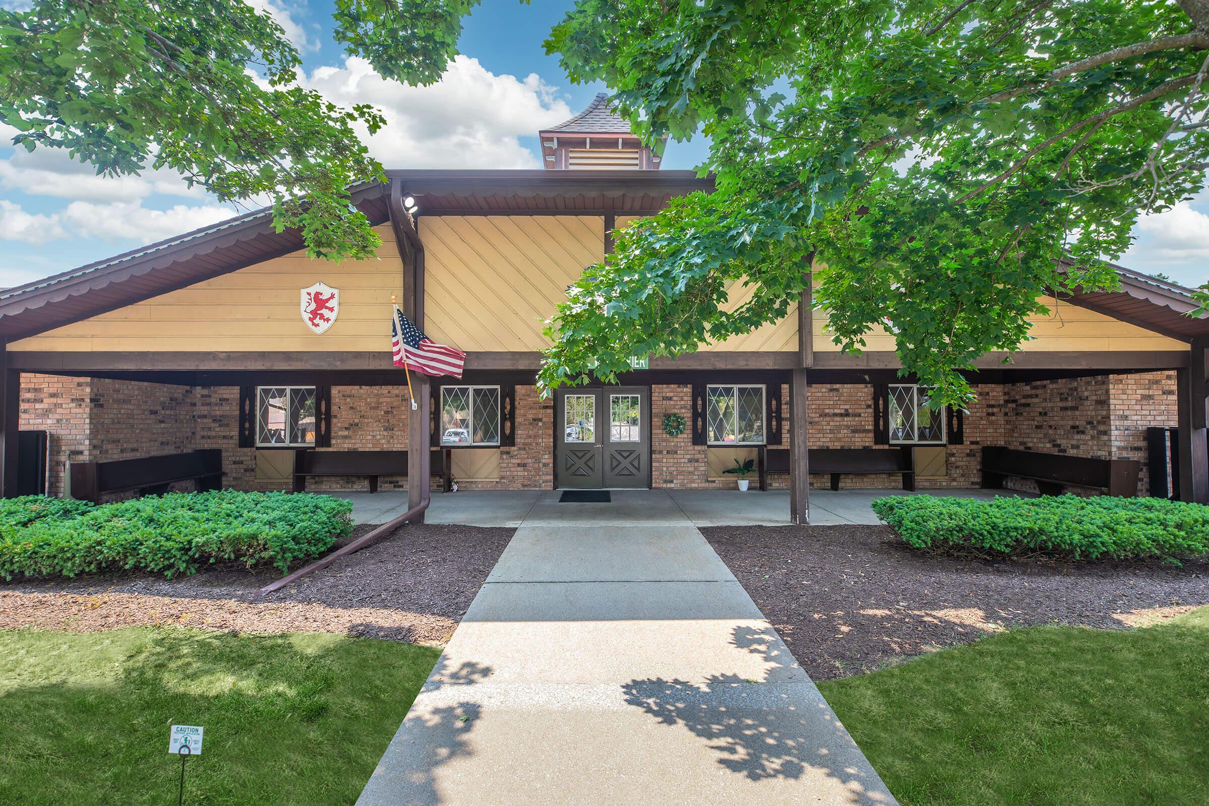 A welcoming building with a large porch, surrounded by green trees and well-maintained landscaping. The exterior features a mix of brick and wooden panels, with a decorative emblem visible. An American flag is prominently displayed, and there are benches along the porch, creating an inviting atmosphere.
