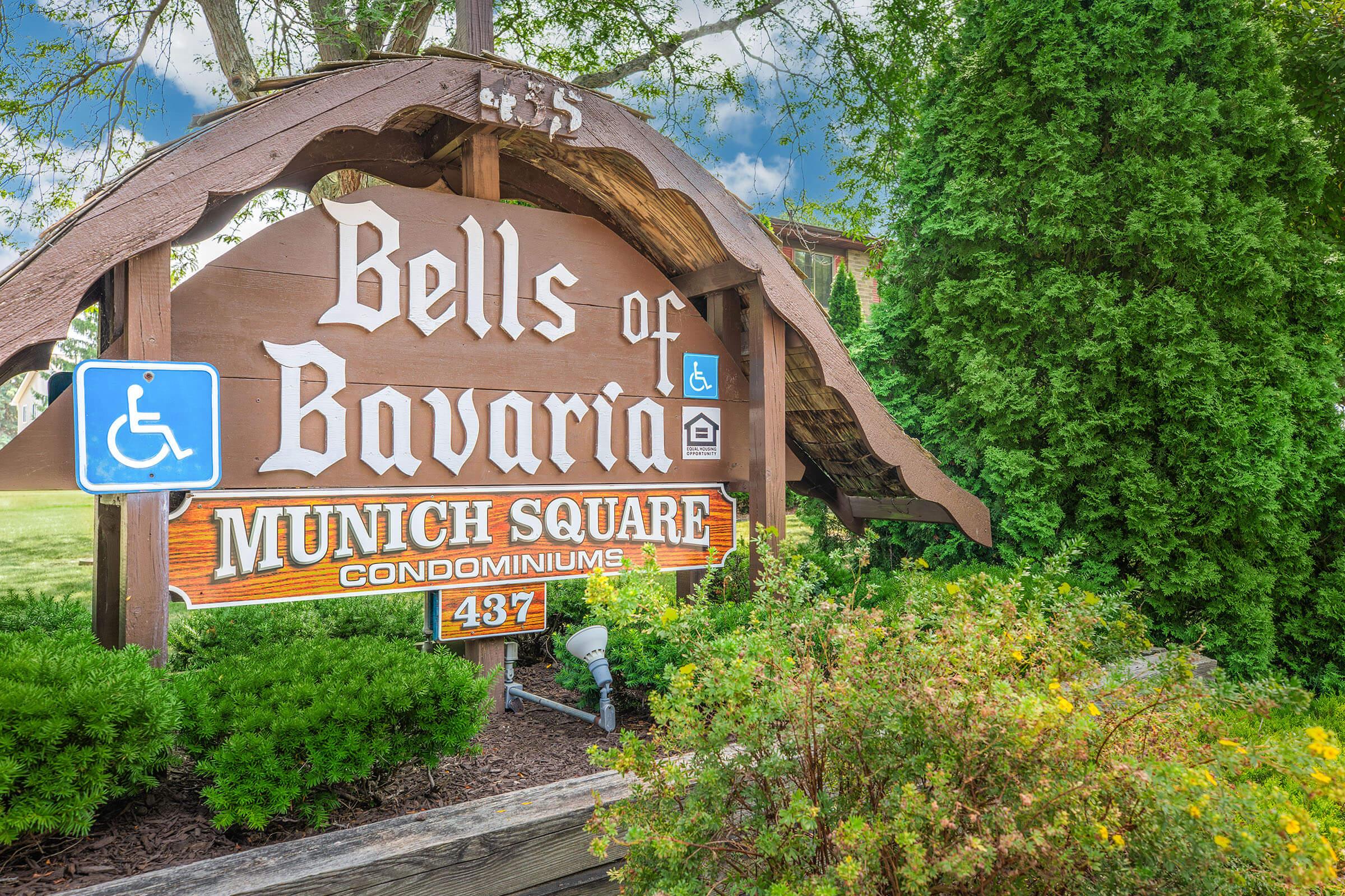 Sign for "Bells of Bavaria," located in Munich Square, indicating the address 437. The wooden sign features colorful lettering and includes a wheelchair accessibility symbol. The background shows green foliage and a blue sky.