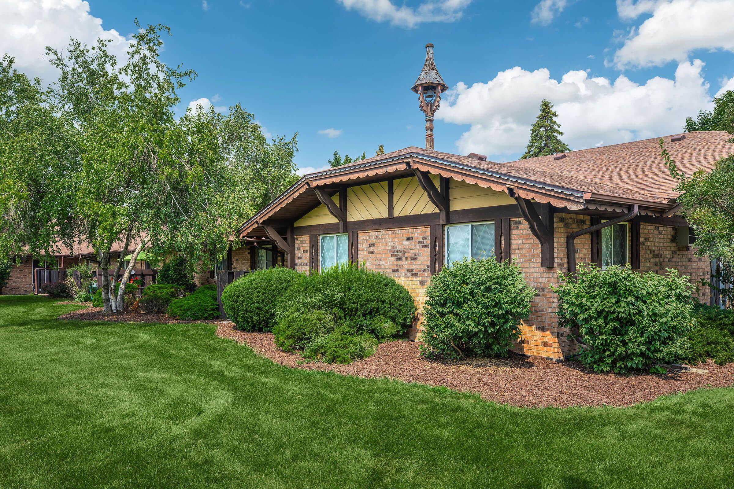 A scenic view of a landscaped home featuring a brick exterior with wooden accents. Lush green bushes and neatly trimmed grass surround the house, under a bright blue sky with fluffy clouds. A decorative feature on the roof adds character to the architectural design.