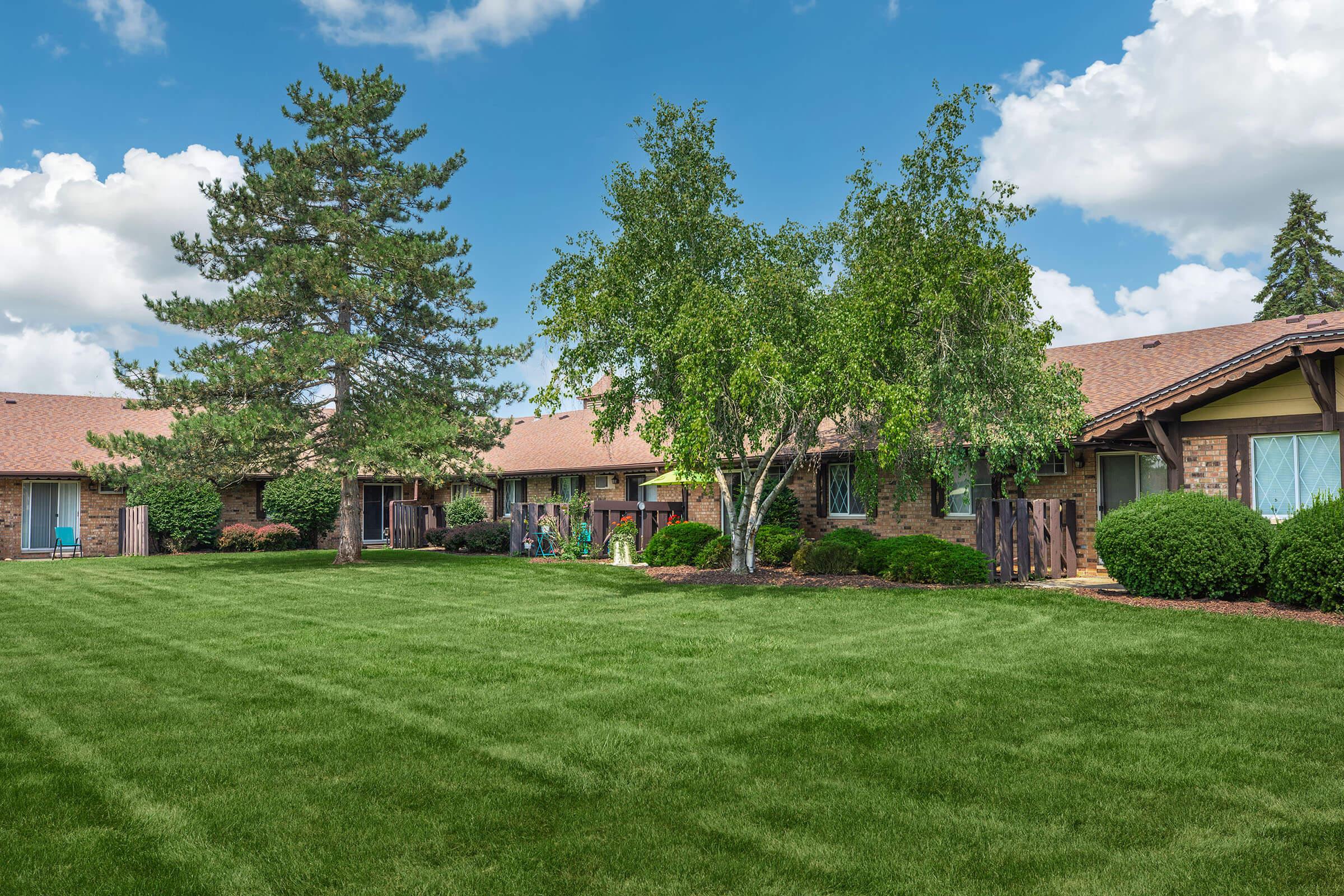 A well-maintained courtyard featuring low-rise buildings surrounded by lush green lawns, trees, and shrubs under a bright blue sky with some fluffy clouds. The area is peaceful and inviting, ideal for relaxation or outdoor activities.