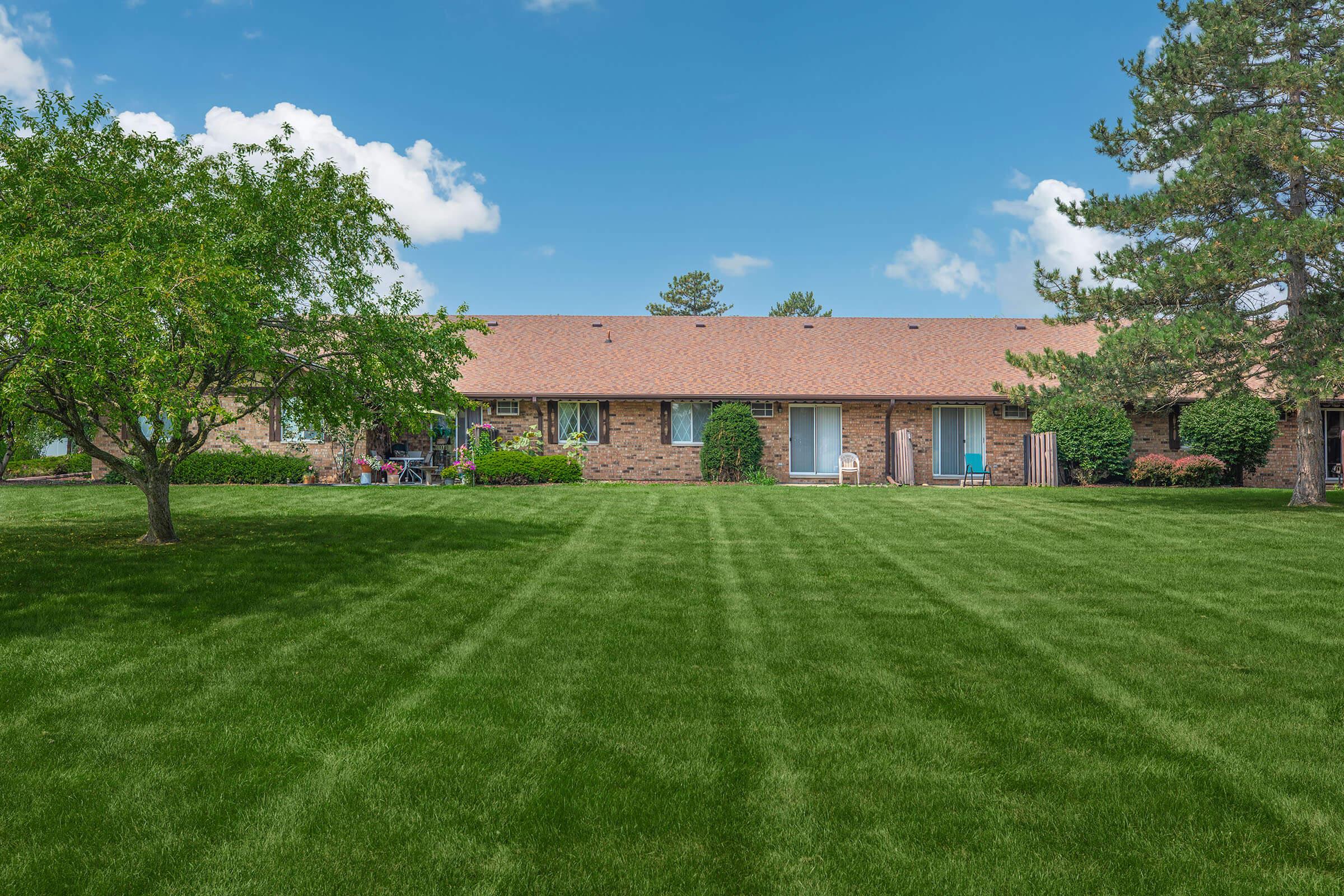 A well-maintained lawn with neatly trimmed grass in front of a row of brick residential buildings. The buildings feature multiple windows and doors, surrounded by trees and shrubs under a clear blue sky with a few clouds.