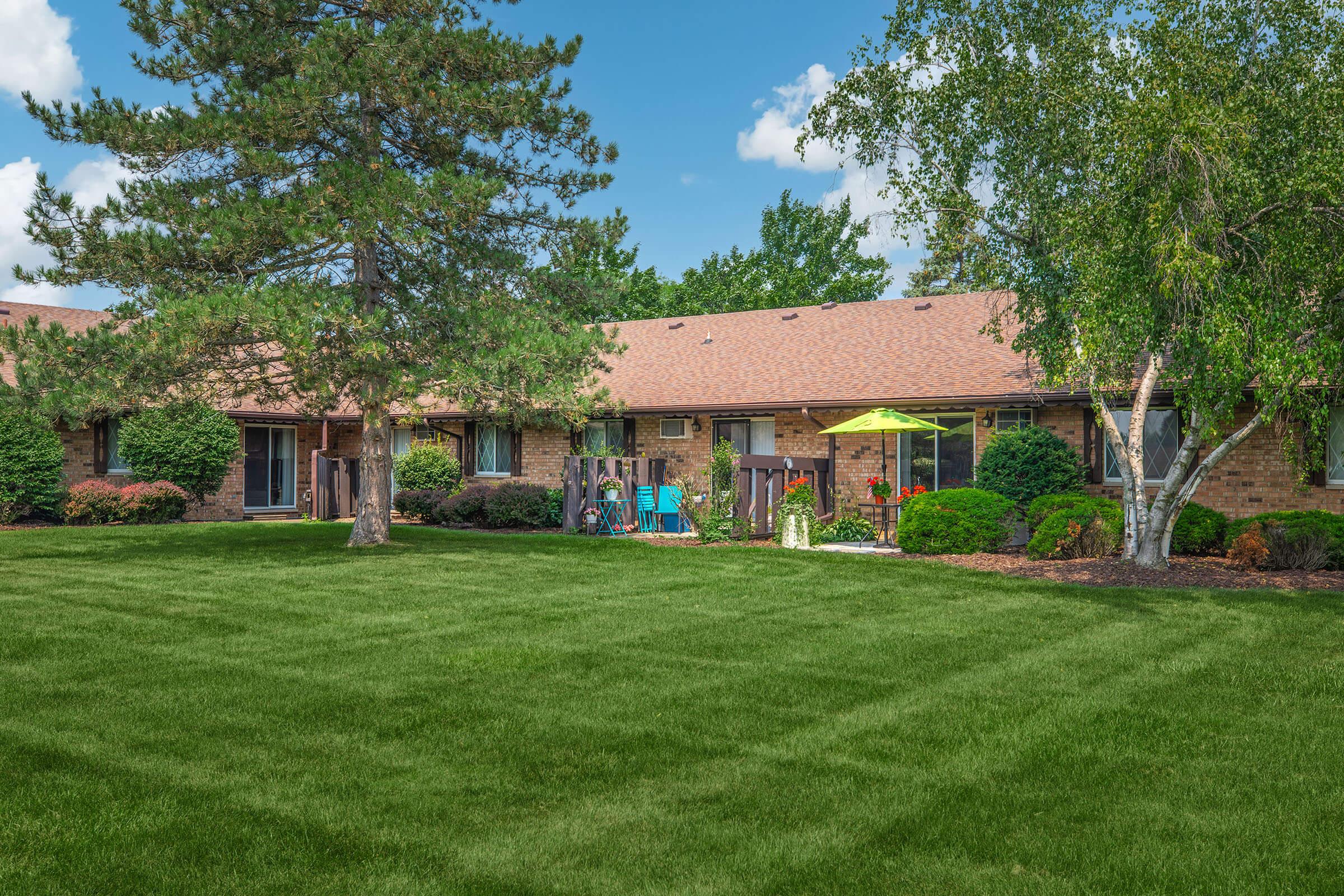 A peaceful residential scene featuring a well-maintained lawn and a row of brick apartments. Trees and shrubs frame the area, and a small patio with colorful furniture and an umbrella can be seen, suggesting an inviting outdoor space for relaxation. The sky is clear with a few clouds.