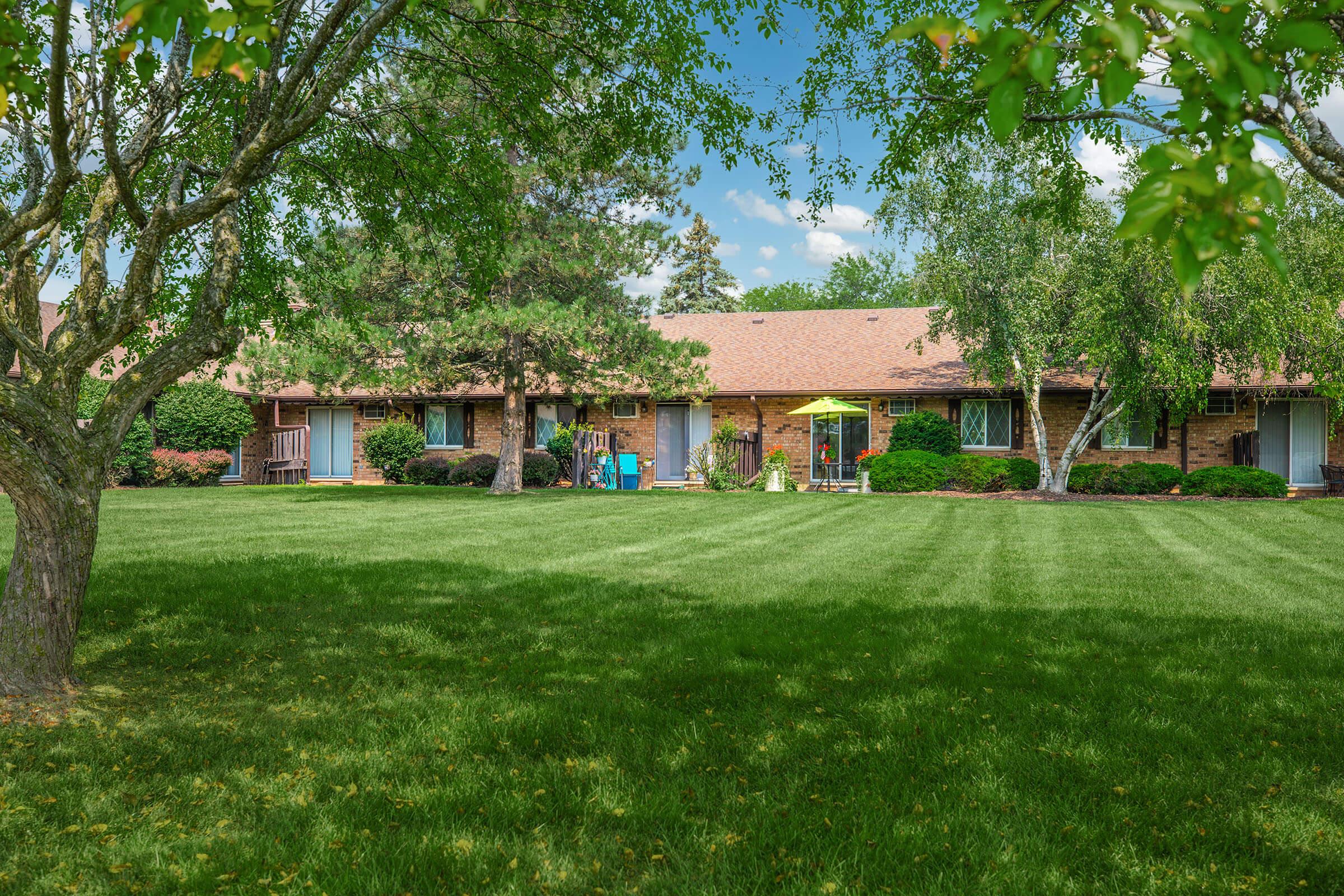 A well-maintained grassy lawn in front of a row of single-story brick buildings with porches. Green trees border the area, and a bright yellow umbrella is visible on the patio. The sky is partly cloudy, creating a serene and inviting atmosphere.