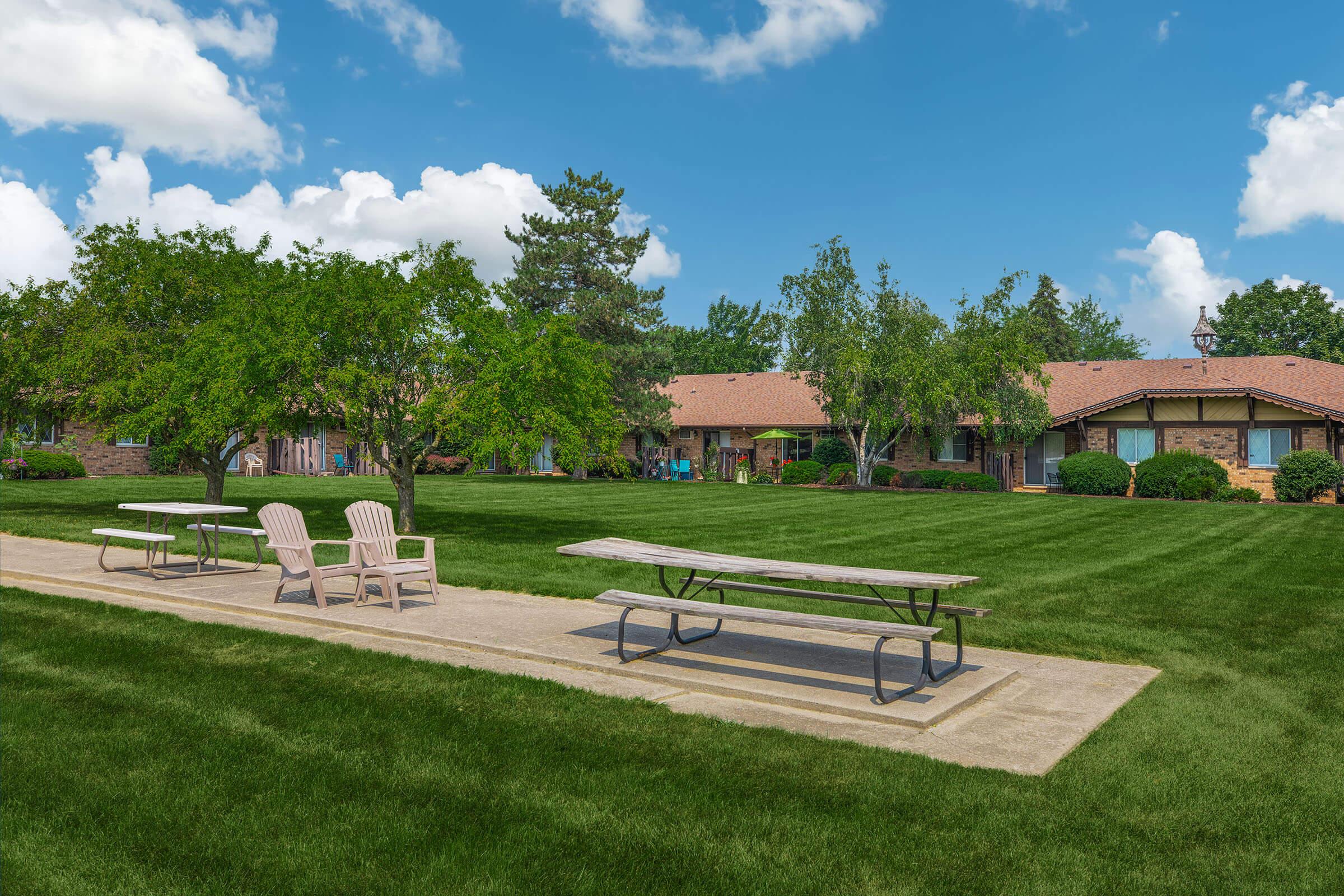 A well-maintained park area featuring two picnic tables and several chairs on a paved path, surrounded by lush green grass. In the background, there are trees and buildings with brown roofs, under a bright blue sky with fluffy white clouds. The scene suggests a peaceful, community-friendly environment.