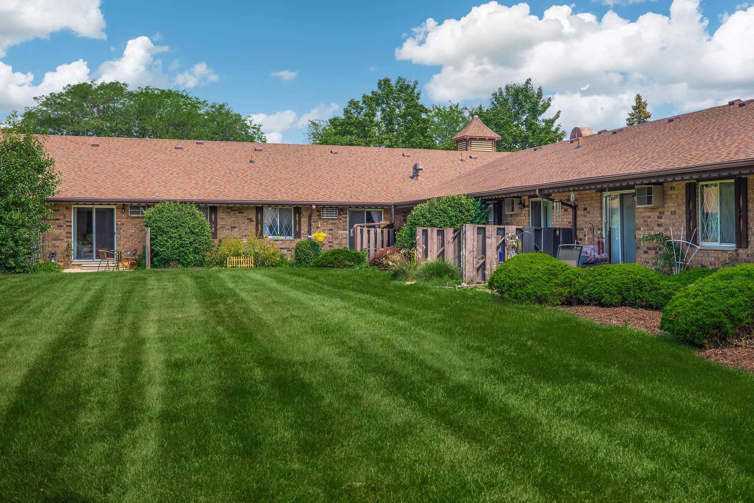 A well-maintained residential backyard featuring a lush green lawn, bordered by neatly trimmed bushes and trees. There are several sliding glass doors leading into the house, and a few outdoor seating areas visible. A blue sky with fluffy white clouds completes the picturesque scene.