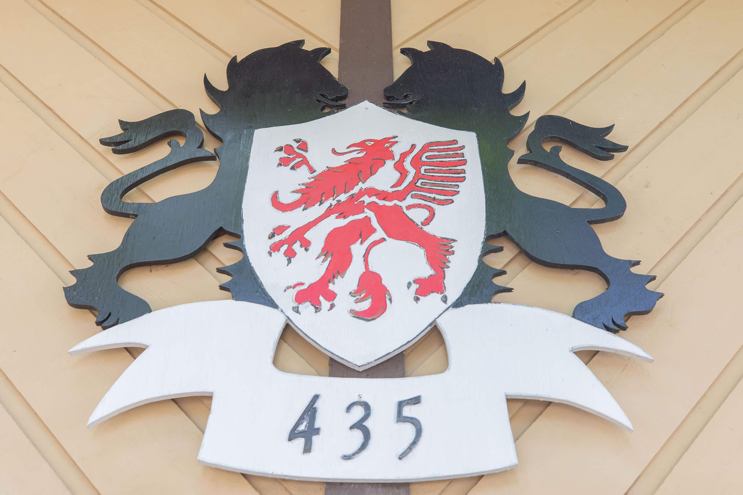 A wooden coat of arms featuring a red lion on a white shield, flanked by two black lions. The design includes a scroll at the bottom displaying the number "435." The overall style is ornate, with intricate detailing in the animals and shield.