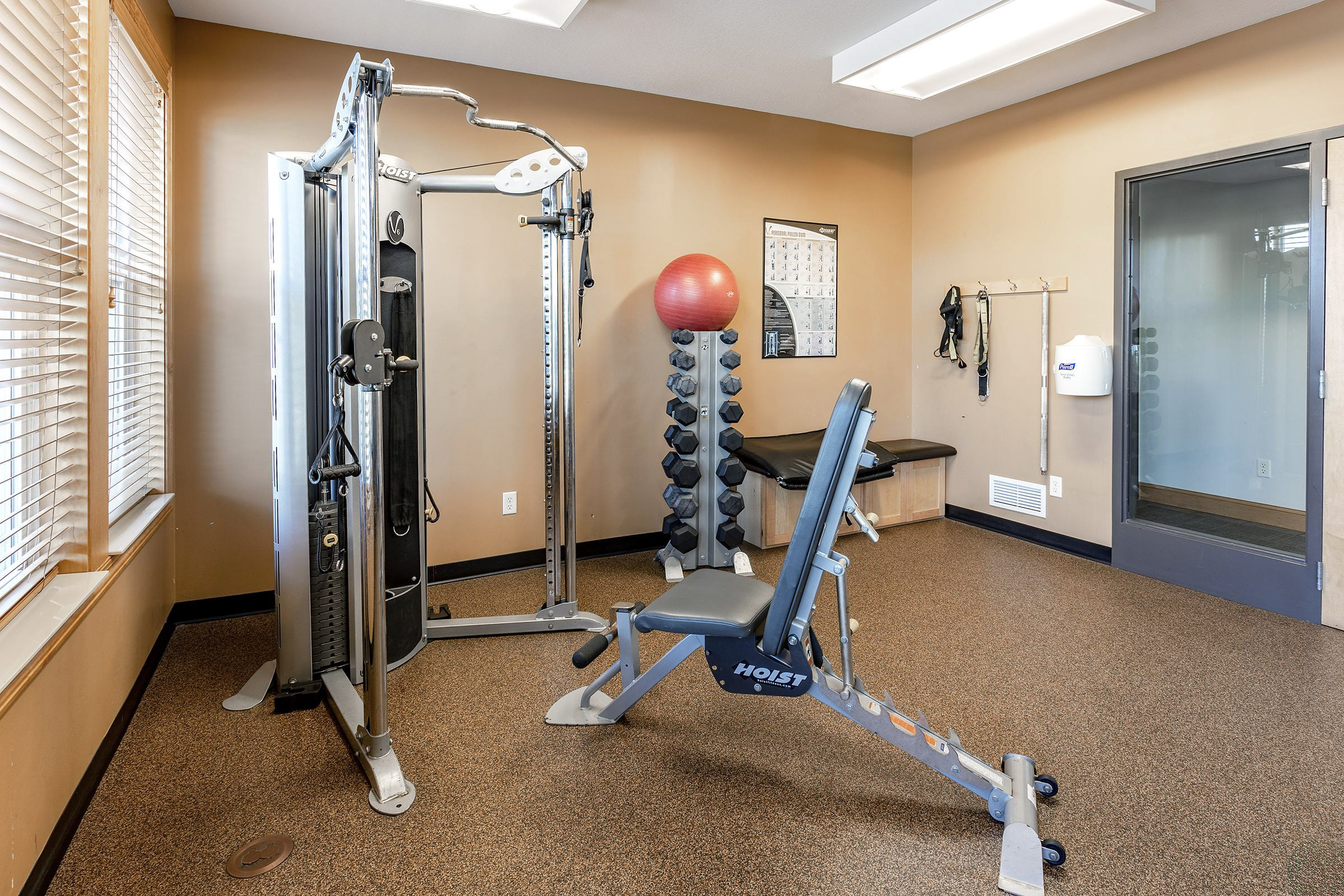 A small fitness room featuring a strength training machine, a stability ball, a weight rack, and a bench. The room has windows allowing natural light, with a beige and brown color scheme, and fitness posters on the wall.
