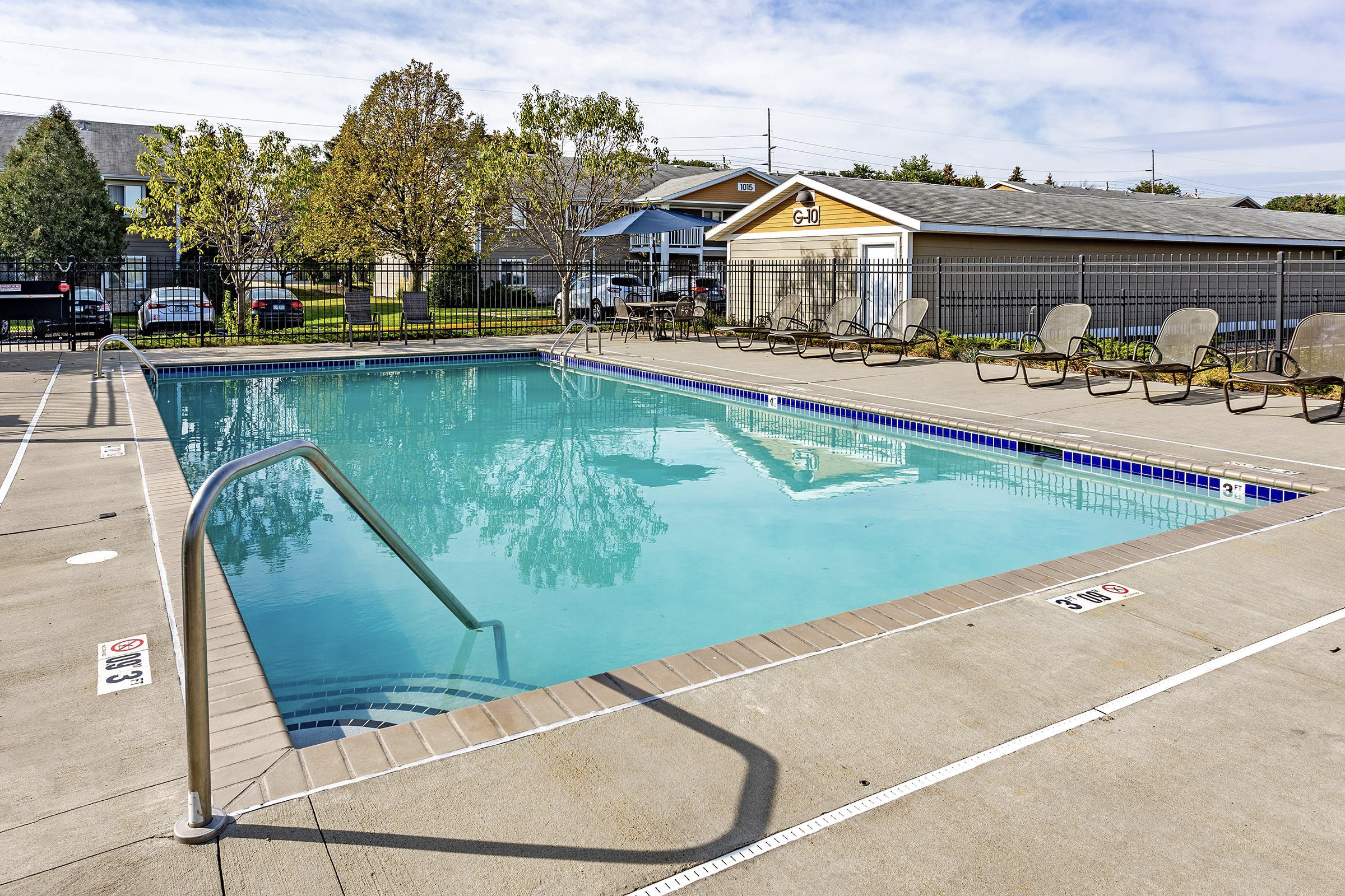 A clear swimming pool surrounded by a concrete deck, lounge chairs, and a fence. Trees and buildings are visible in the background under a blue sky with some clouds.