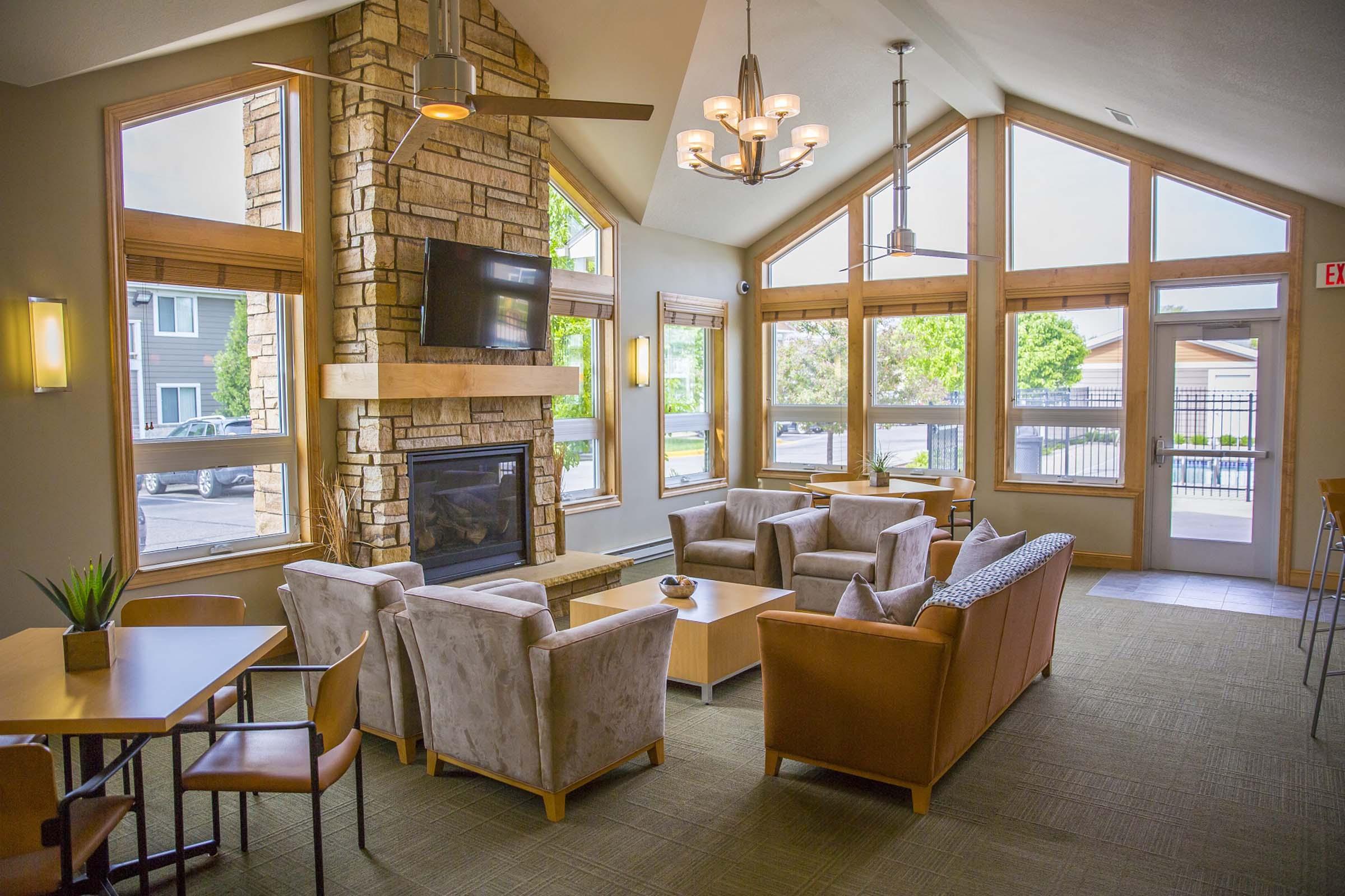A cozy lounge area featuring comfortable seating with light-colored chairs and a sofa, a stone fireplace, and large windows that allow natural light to fill the space. There are wooden accents throughout, along with modern light fixtures and a small table with a plant, creating a welcoming atmosphere.