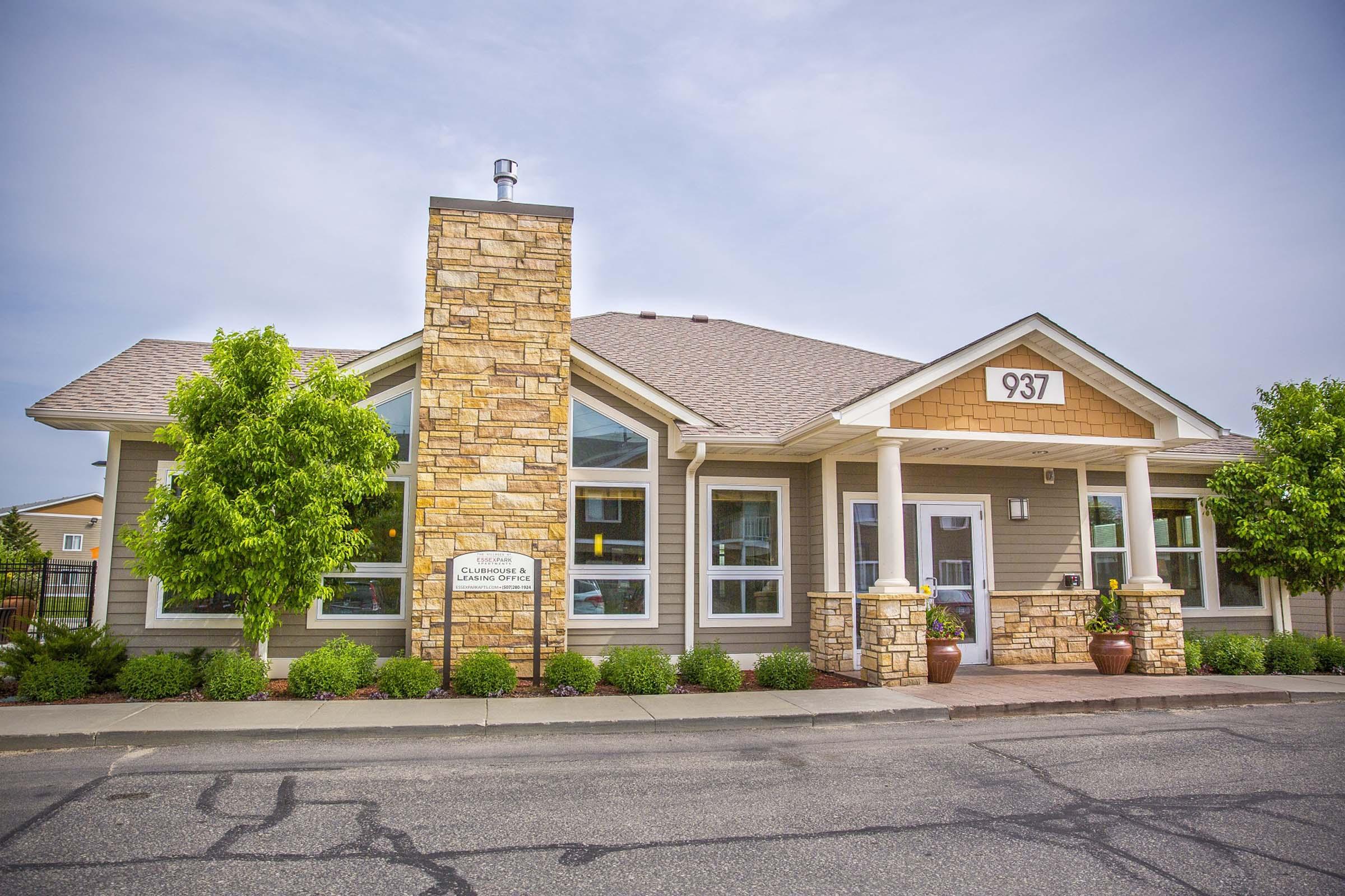 A charming single-story building featuring a stone facade, large windows, and a welcoming entrance. Flanked by neatly trimmed bushes and trees, it has a sloped roof and a prominent address number, 937, displayed above the door. The setting is well-maintained with a paved front area.