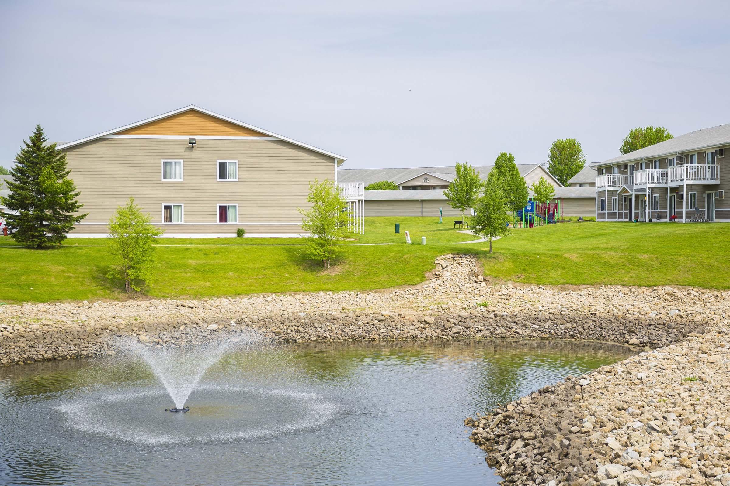 A serene landscape featuring a tranquil pond with a fountain in the foreground. In the background, there are two residential buildings surrounded by lush green grass and trees, creating a peaceful suburban environment.