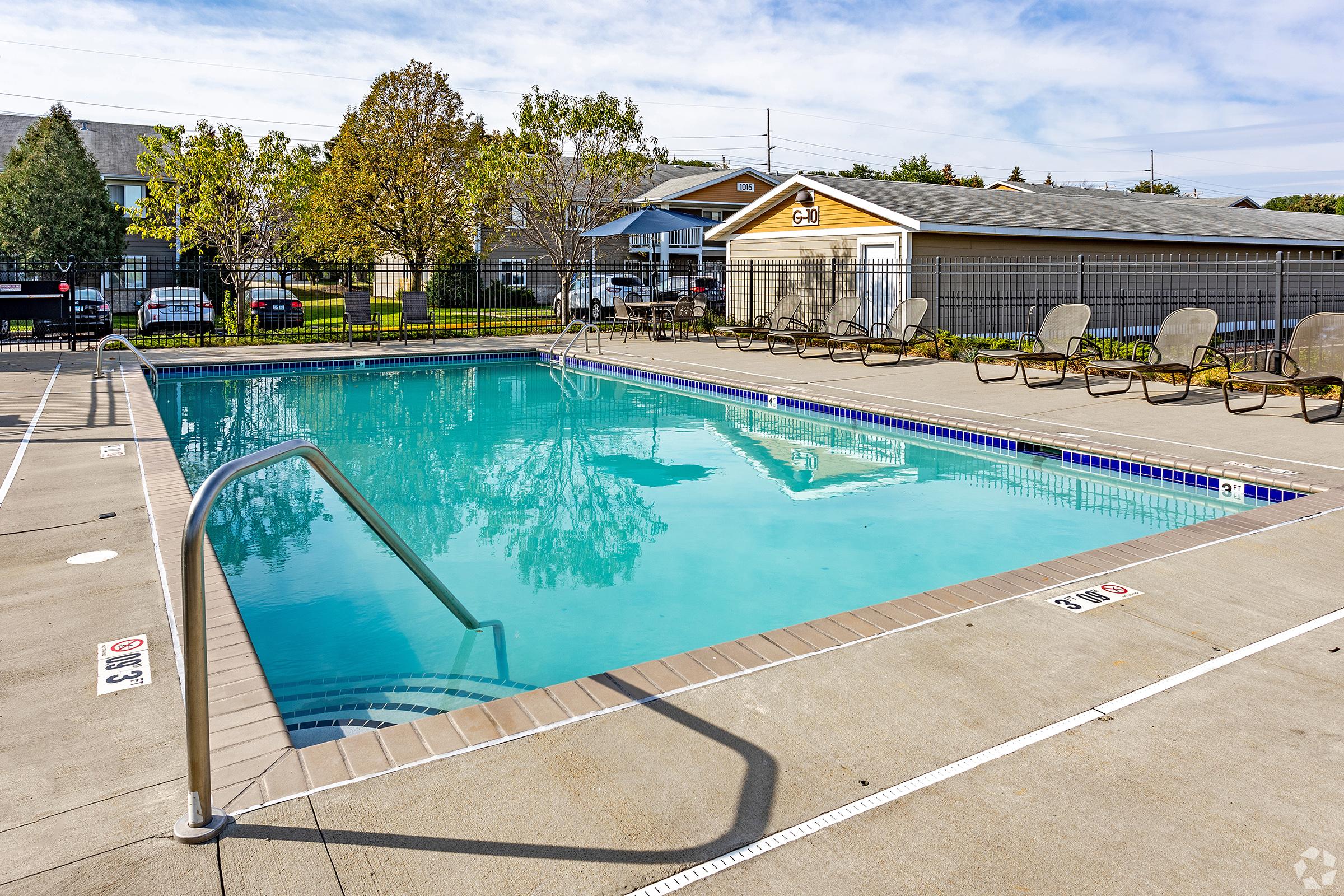 A clear swimming pool surrounded by a concrete deck, with lounge chairs positioned around the perimeter. The pool has a railing on one side and is situated in a well-maintained outdoor area, featuring trees and buildings in the background. Bright blue skies above enhance the inviting atmosphere.
