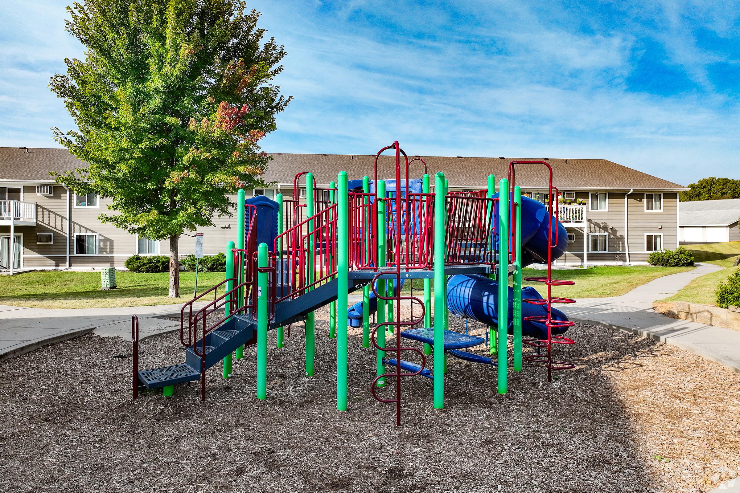 A brightly colored playground structure with green, blue, and red components, including slides, climbing bars, and ladders. Surrounding the playground are grassy areas and residential buildings in the background, under a clear blue sky. Wood chips cover the ground beneath the play equipment.