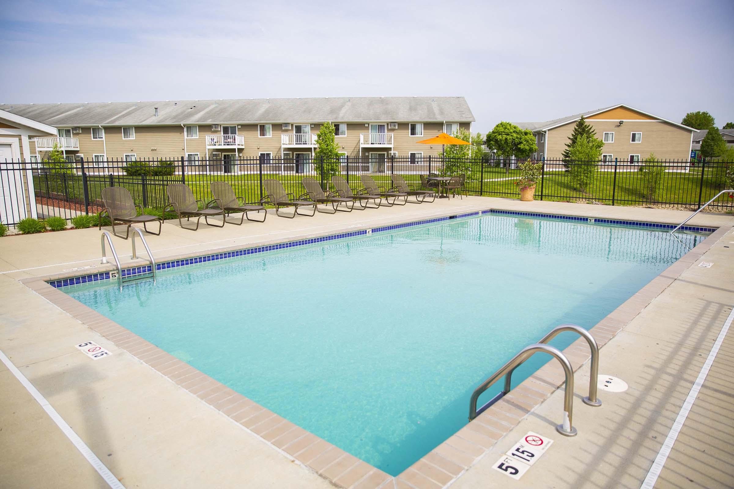 A clear blue swimming pool surrounded by a patio area with lounge chairs. In the background, there are residential buildings with multiple units, green grass, and trees. The scene is well-lit and inviting, suggesting a recreational space for relaxation and leisure.
